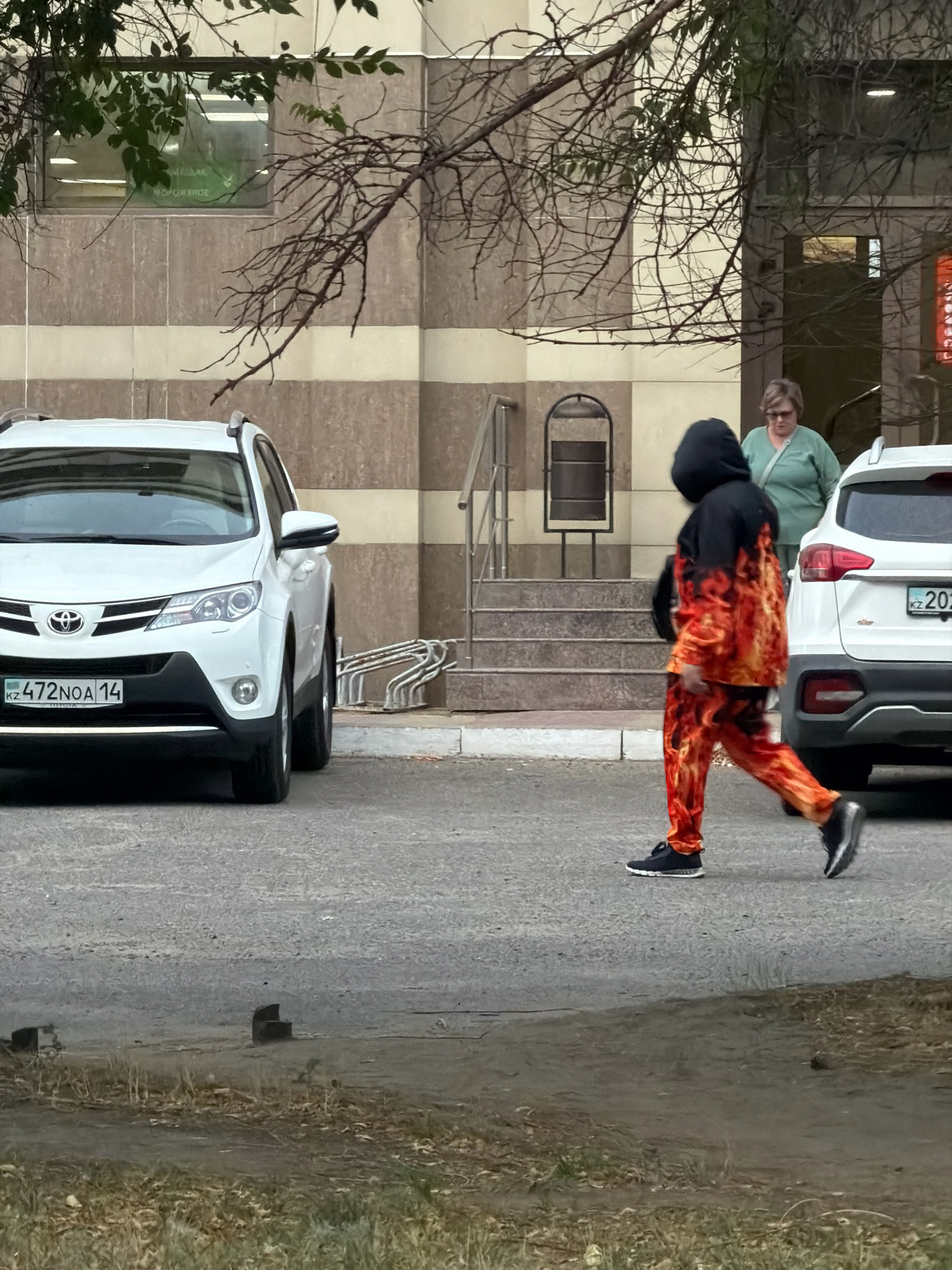 The image depicts an outdoor scene in the city of Pavlodar, Kazakhstan. In the foreground, a person wearing a black hoodie and pants with a flame pattern walks from right to left across a paved area. The person is wearing black sneakers. Behind this person, a woman in a light green shirt stands near a white SUV. To the left, a white Toyota SUV is parked. The background features a building with a beige and brown striped facade, and a tree branch with green leaves extends into the frame from the upper left. The ground in the foreground is covered with dry grass and dirt. A license plate on the white Toyota SUV reads "KZ 472NOA 14". Another license plate on the white SUV to the right partially visible shows "KZ 202". A bicycle rack is visible near the entrance of the building.