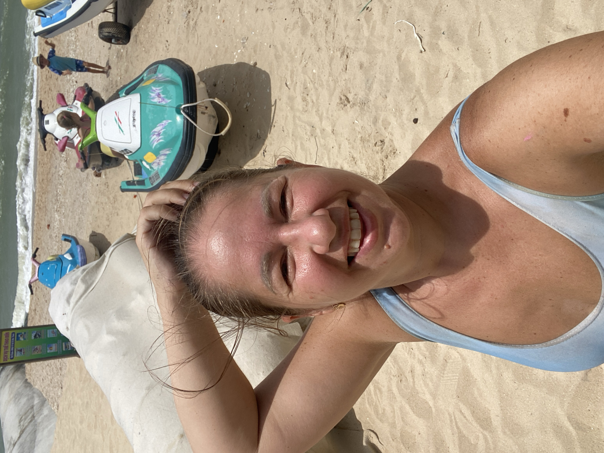 In Mũi Né, Vietnam, on a sandy beach, a woman is taking a selfie. She is smiling and has her arm raised, with her hair partially obscuring her face. She is wearing a light blue swimsuit. In the background, several jet skis are parked on the sand, with the ocean and waves visible beyond them. A young boy wearing a straw hat and swim trunks is standing near one of the jet skis, and a large white object, possibly a beanbag chair, is also in the frame. A sign with Vietnamese text is present near the beach's edge.