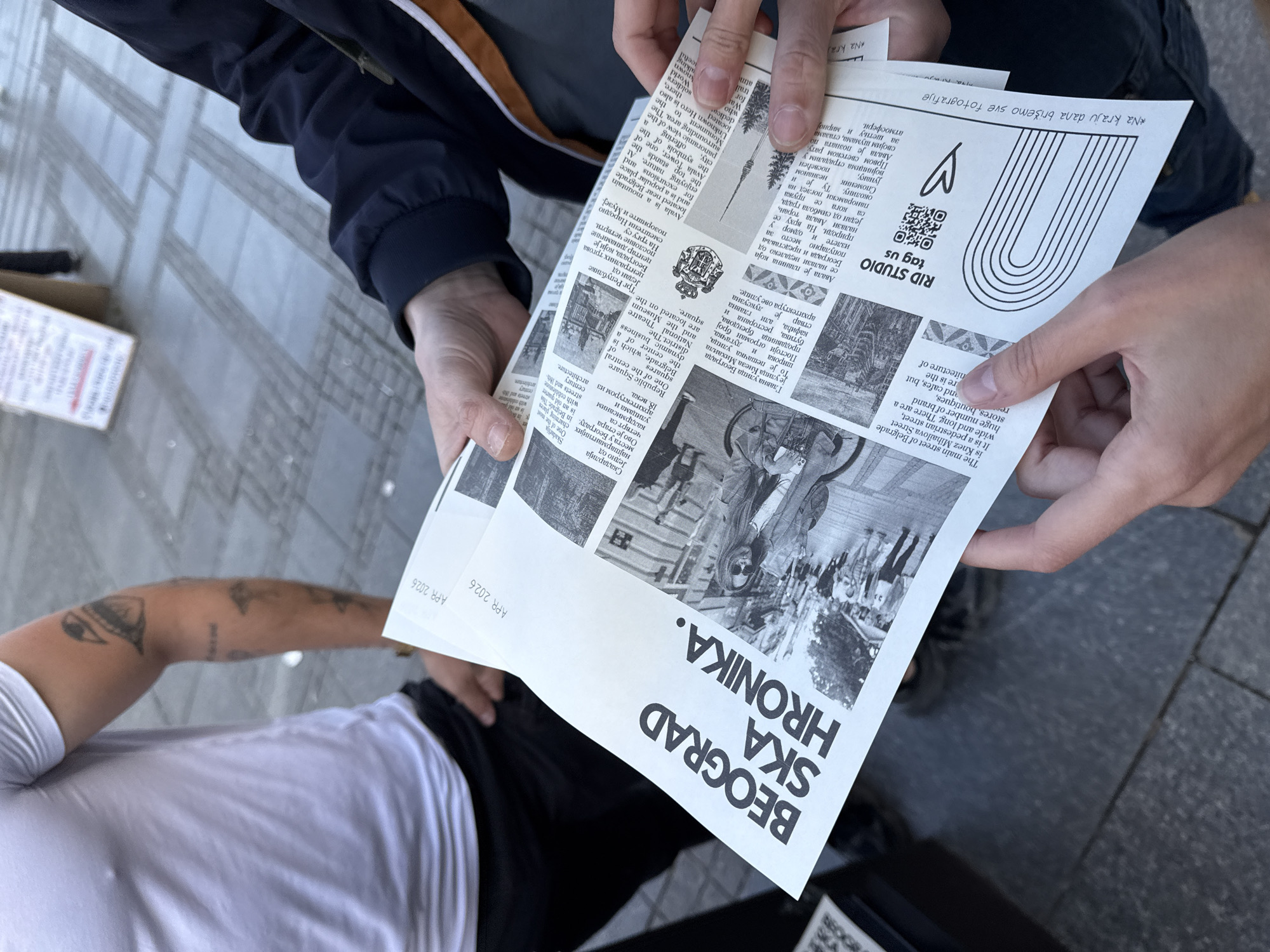 The image shows a close-up, overhead view of two individuals interacting with a folded newspaper or magazine. One person, wearing a dark blue jacket with a white and orange stripe on the sleeve, holds the publication open with both hands, presenting it towards the viewer. The other person, partially visible in the lower-left, wears a white t-shirt, and a tattooed arm is prominent. This second person appears to be receiving or jointly viewing the paper.
The publication is black and white, prominently displaying the title "BEOGRAD SKA HRONIKA." (Belgrade Ska Chronicle) in bold, sans-serif text. The date "APR 2026" is visible in the bottom-left corner of the paper. It features a mix of articles in both English and Serbian (Cyrillic), accompanied by various photographs and graphics. Notable text includes references to Belgrade landmarks such as Knez Mihailova Street, Republic Square, Avala, and Skadarlija, confirming the location context of Belgrade, Serbia. The paper also includes a "RID STUDIO" logo, a QR code, a heart icon, and the phrase "*Na kraju dana brišemo sve fotografije" (At the end of the day, we delete all photos) in the top-right corner.
The setting is an outdoor paved area, likely a sidewalk or plaza, characterized by a grey, patterned surface. A cardboard box is partially visible on the ground to the lower-left. The lighting suggests it is daytime. The activity depicts an informal interaction, possibly an exchange or shared reading of the local publication.