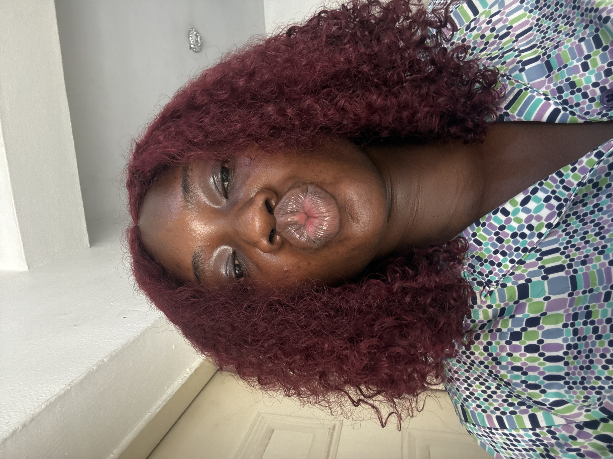 A woman with dark skin and dark red curly hair puckers her lips for the camera. She wears a collared shirt with a geometric pattern of teal, green, purple, and white. The background includes a white wall and door. There is no information in the image to indicate a location in Abuja, Nigeria.