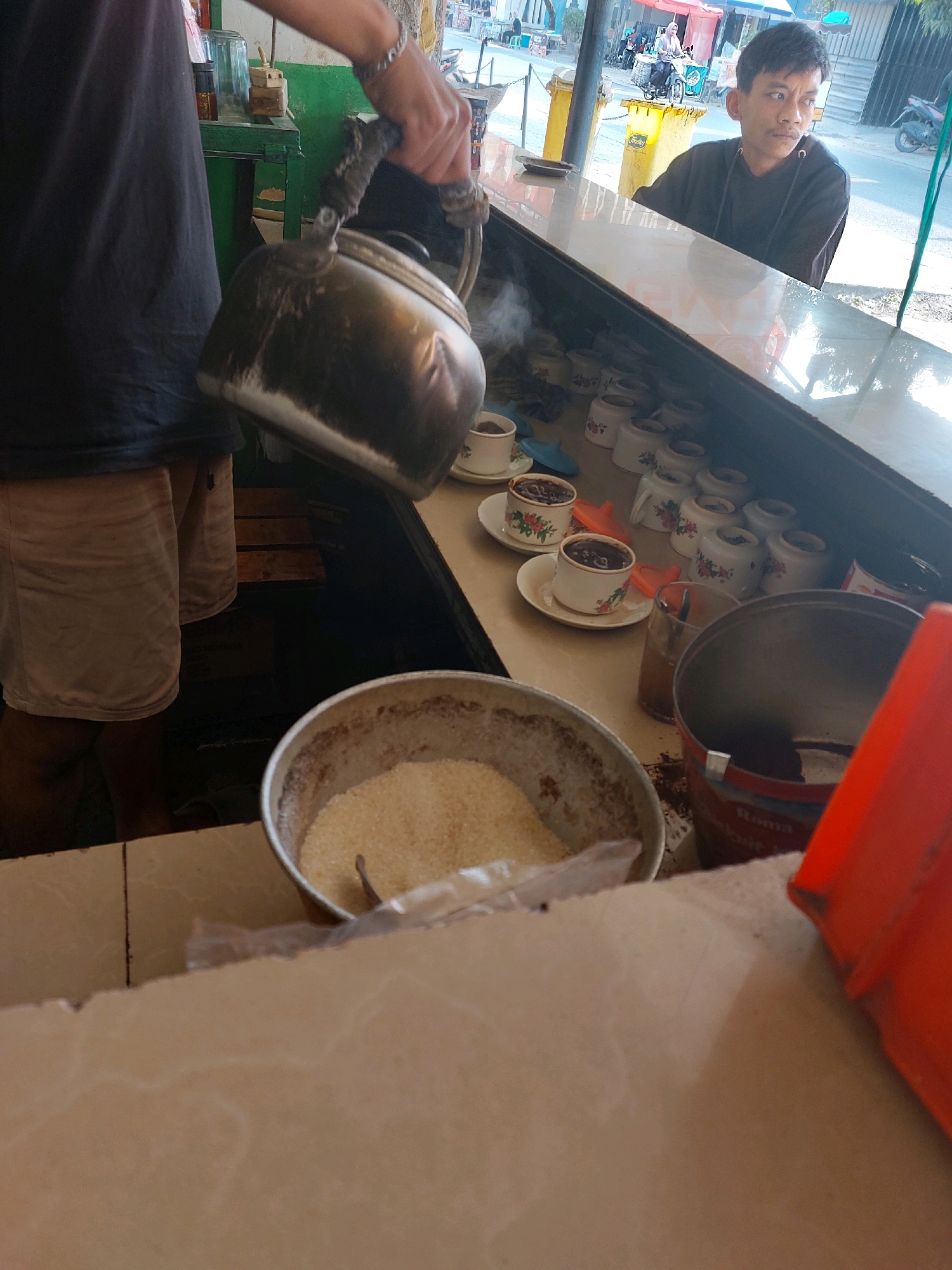 This is a medium shot, eye-level, outdoor, daylight image taken at a roadside stall in Sumuragung, Indonesia. A person wearing a black t-shirt and light-colored shorts is holding a metal kettle, pouring steam into a bowl filled with sugar. In the background, a man with dark hair is seated at a counter, looking to the side. The counter is lined with small cups and saucers. Various items, including a glass of liquid and a plastic container, are visible on the counter. The environment appears to be a busy street with some buildings and vehicles in the distance.