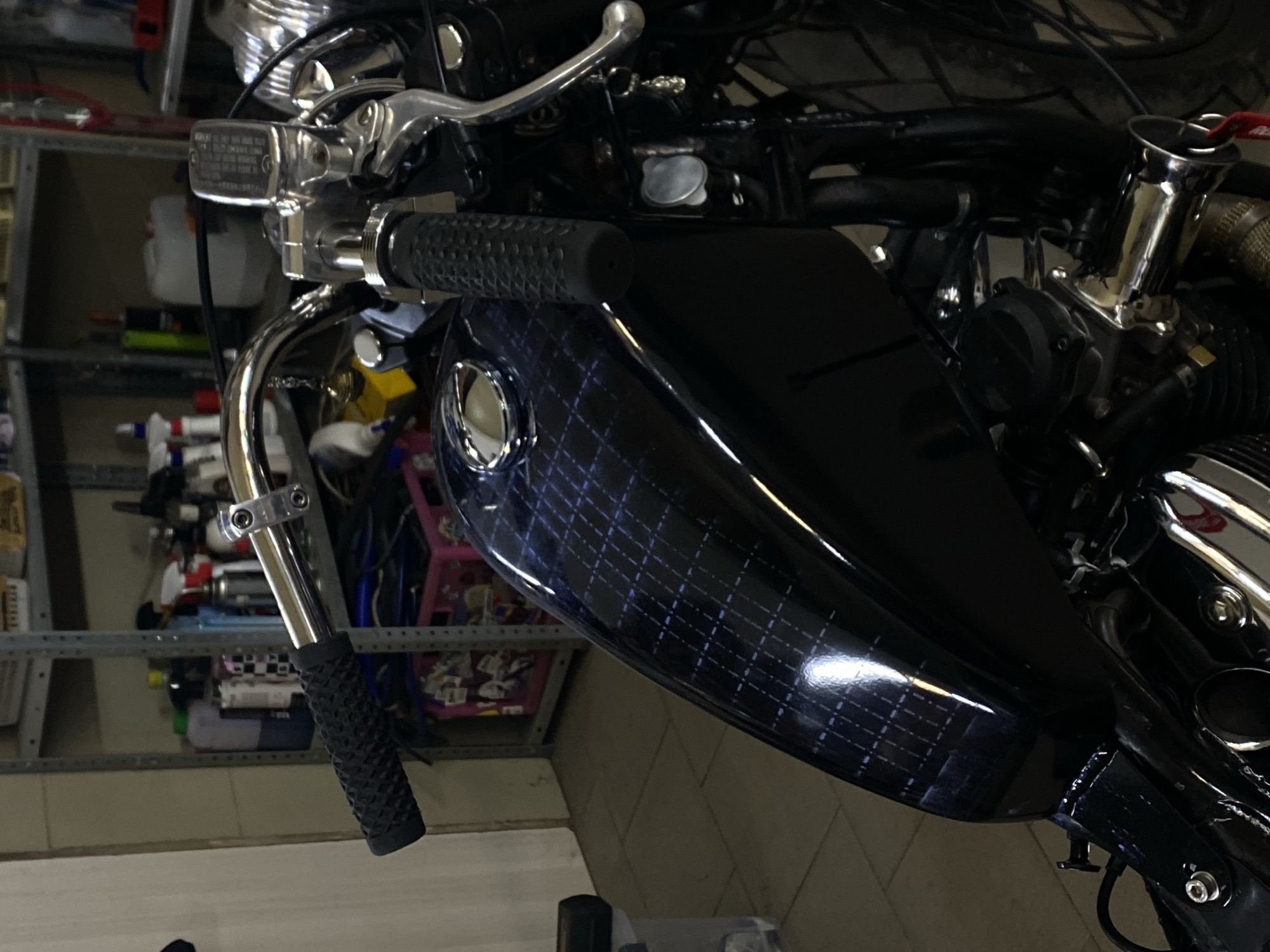 The image shows a close-up view of a black motorcycle's fuel tank and handlebars, set in a dimly lit garage environment. The fuel tank features a dark, glossy finish with a subtle grid pattern in a lighter shade, possibly a custom paint job or decal. The handlebars are chrome, with black textured grips and a visible brake fluid reservoir. A small decorative charm hangs from the handlebar. In the background, shelves filled with various bottles and containers, along with a pink plastic bin, suggest storage of supplies. The flooring appears to be tiled. The image does not contain any information that would allow for its localization to the city of Agronom, Russia.