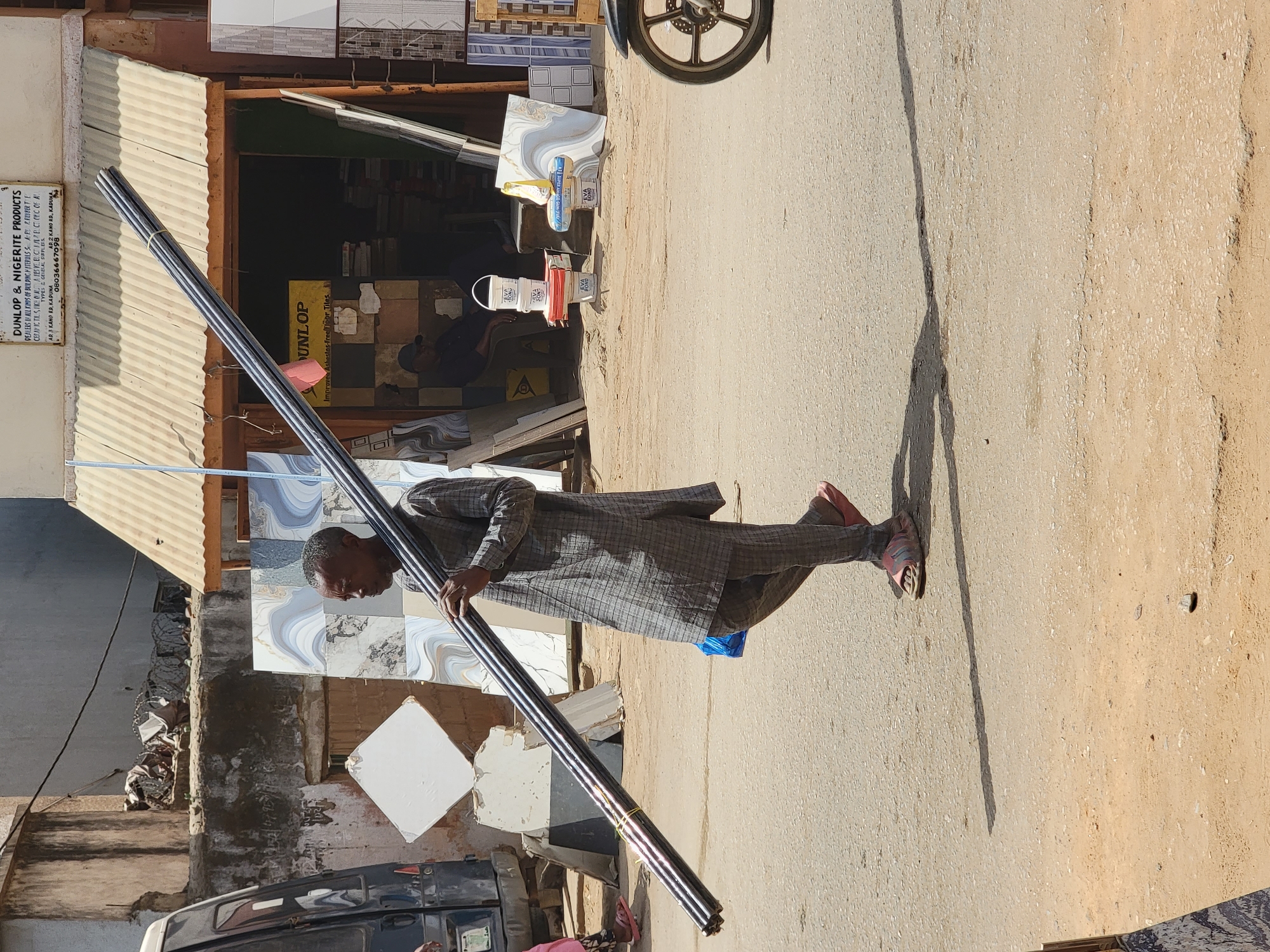 A man in a gray plaid robe walks on a paved road carrying a bundle of dark, glossy rods. He is walking away from a building which appears to be a shop. The shop sells tiles and materials, as indicated by the display of tiles and a sign that reads "DUNLOP & NIGERITE PRODUCTS, DEALERS IN ALL KINDS OF BUILDING MATERIALS". A motorcycle is parked near the shop. The location, identified from the shop sign, is at 3 Kano Road, Kaduna, suggesting this is in Nigeria, possibly in the city of Maiburiji, as requested.