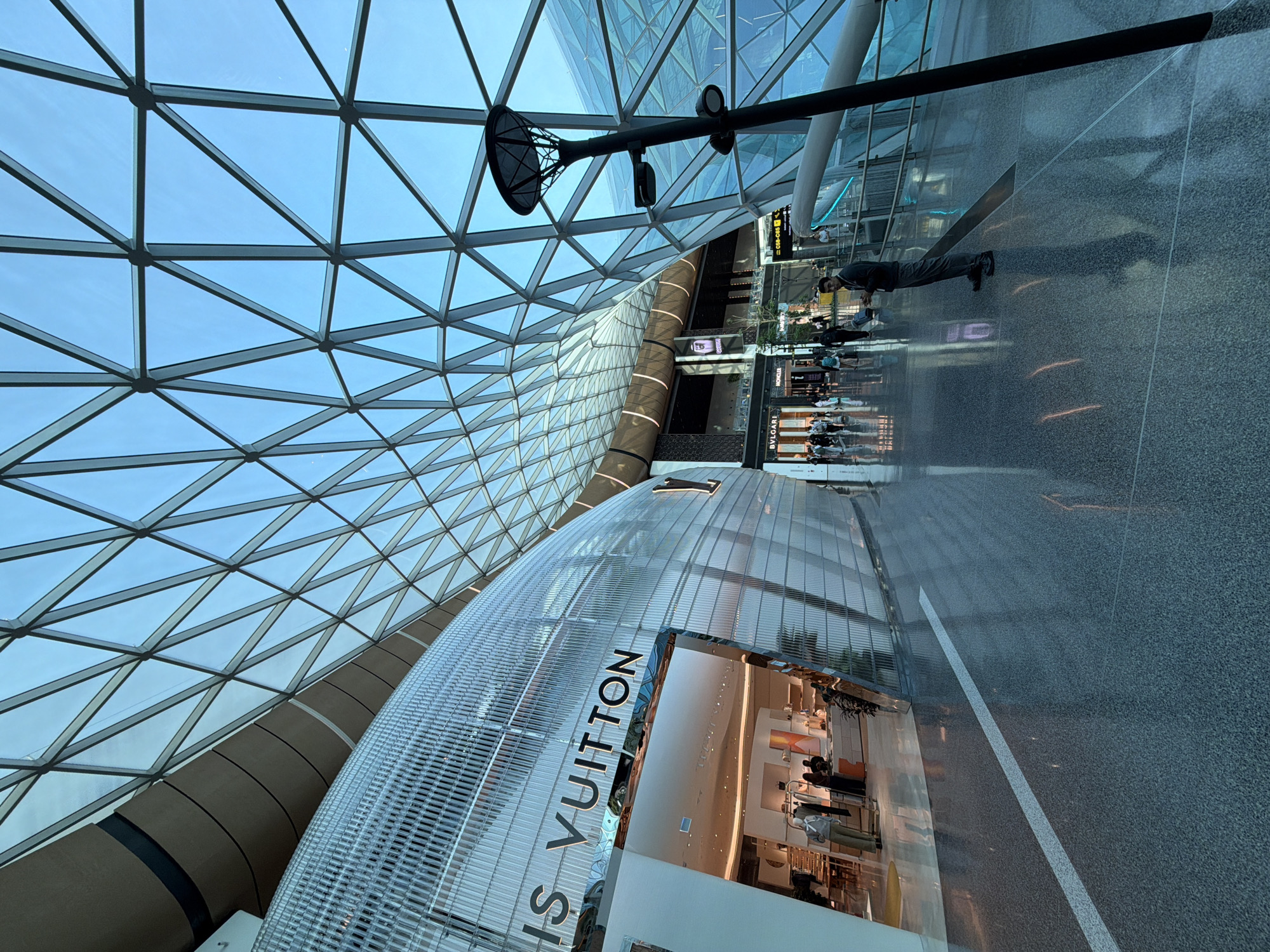 The image depicts an interior view of Hamad International Airport in Doha, Qatar, characterized by its expansive, geometrically patterned glass and steel ceiling. Prominently featured on the left is the facade of a Louis Vuitton store, its curved, translucent structure illuminated from within. Inside the store, mannequins display merchandise, and several individuals are visible browsing.
To the right of the Louis Vuitton store, a dark lamppost with multiple light fixtures stands tall. Further down the walkway, other retail establishments like Bvlgari and Moncler are visible, with shoppers and travelers moving about. The polished, speckled flooring reflects the ambient light and the architectural elements of the space. A man in a dark jacket and grey pants is standing near the center of the frame, looking towards the left. The overall environment is modern and luxurious, typical of a high-end international airport terminal. The lighting suggests it is either early morning or late afternoon, with natural light filtering through the glass ceiling.