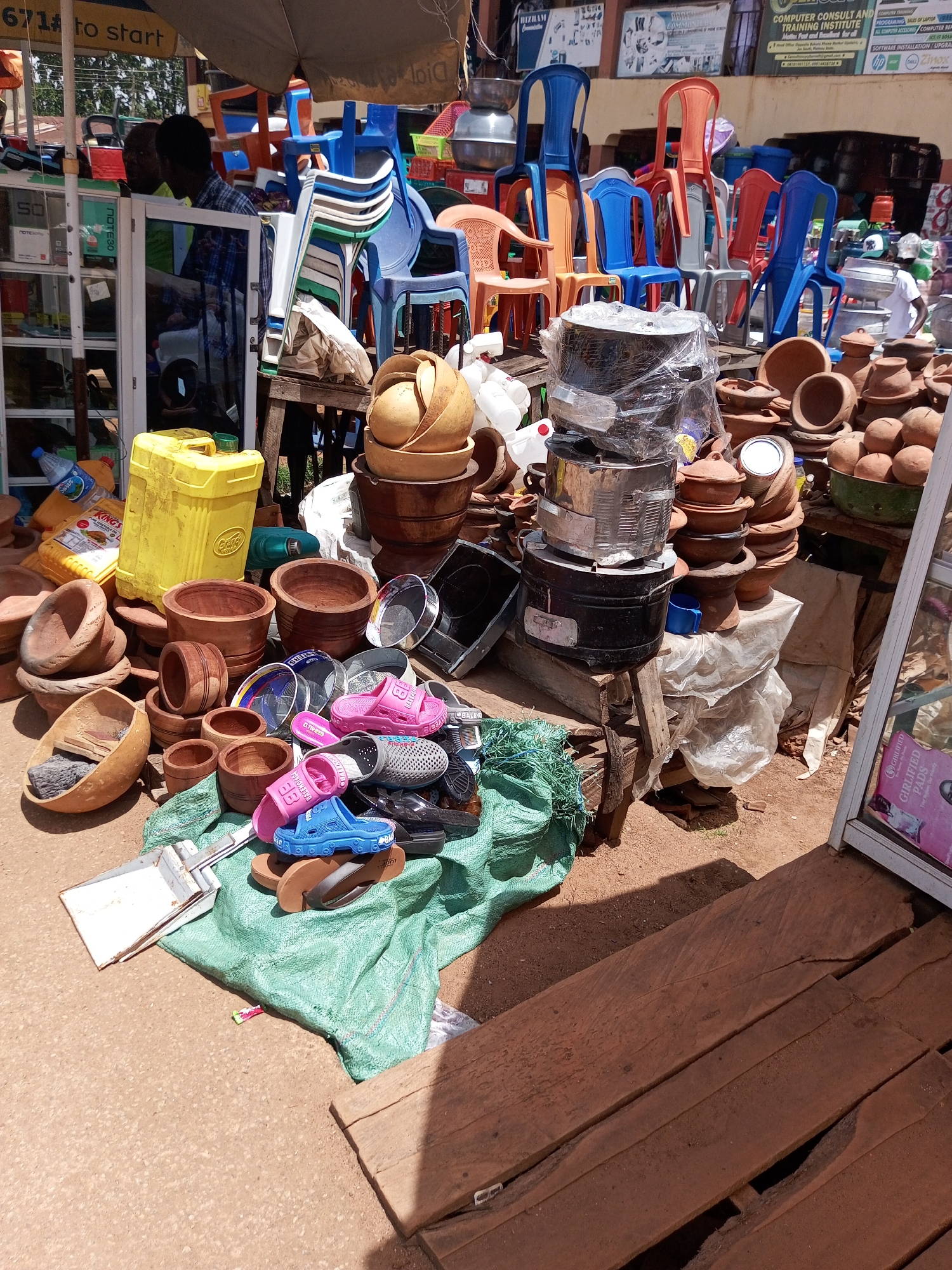 The image depicts an outdoor market scene in Bukuru, Nigeria. In the foreground, a collection of clay pots and bowls are arranged on the ground, some stacked on top of each other. Several pairs of colorful plastic sandals, in pink and blue, are placed on top of a large green tarp. To the left, a yellow plastic container and a white dustpan are visible. In the middle ground, various chairs, including blue, orange, and red plastic chairs, are stacked and arranged. A man in a blue and white checkered shirt is partially visible in the background, standing near a shelf unit. The environment appears to be a busy marketplace with merchandise displayed on the ground and on shelves.