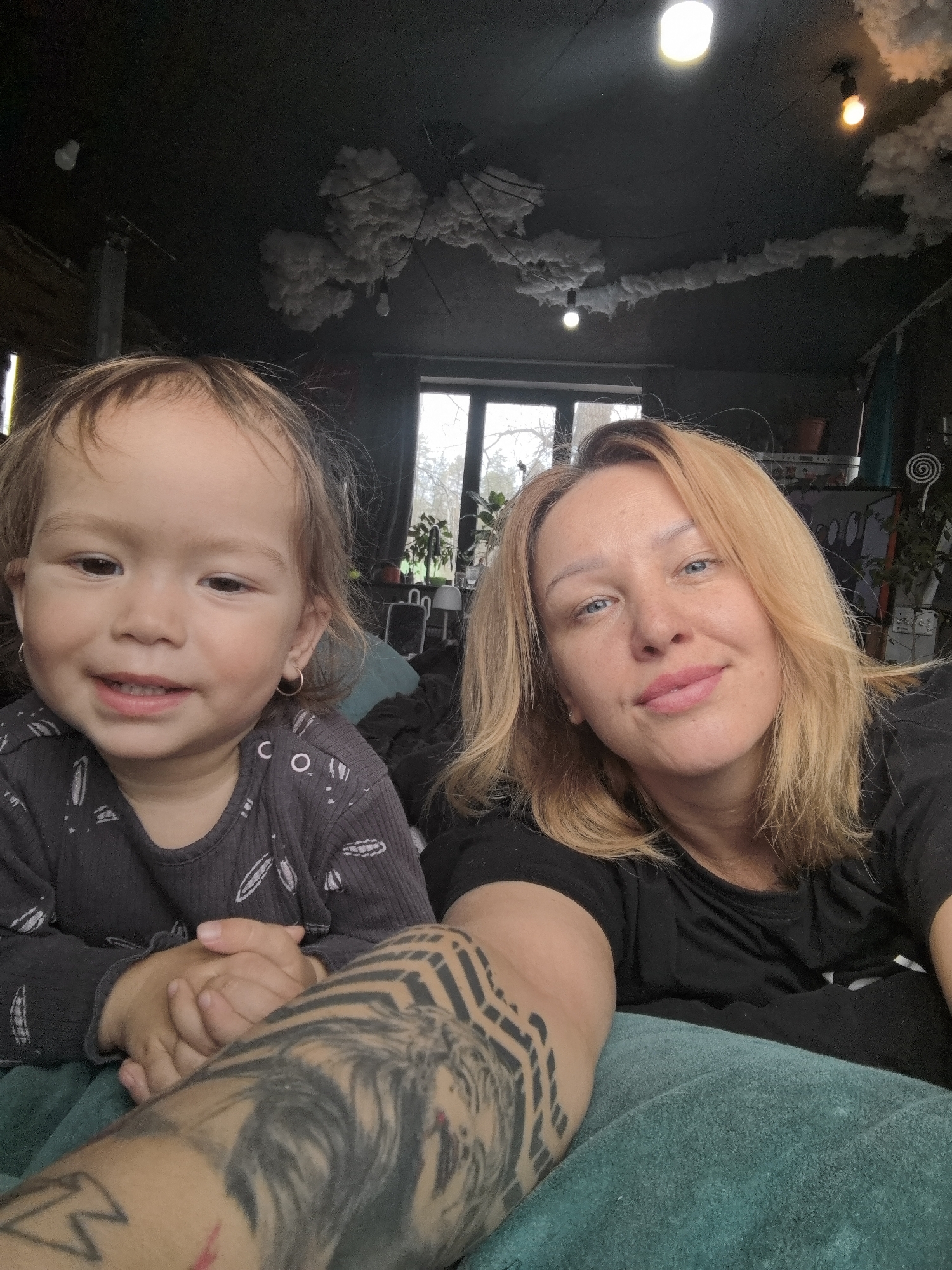 A selfie-style photograph shows a woman and a young child indoors. The child is in the foreground to the left, smiling slightly with their hands clasped. The woman is to the right, also smiling gently. The woman has blonde hair and is wearing a black shirt. A portion of the woman's arm with a tattoo is visible in the foreground. The background reveals a room with a window, indoor plants, and decorative cloud-like installations on the ceiling. The photograph does not include information to verify the location as Noviy, Russia.