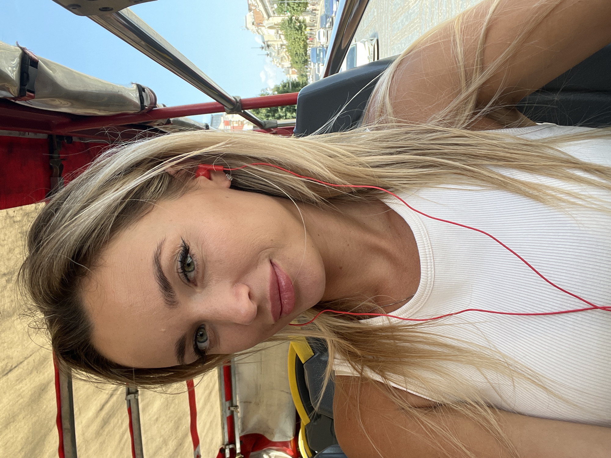 A young Caucasian woman with blonde hair and green eyes is captured in a selfie. She is wearing a white ribbed tank top and red earphones. The woman is seated in what appears to be an open-top tour vehicle, possibly a tuk-tuk, with a tan fabric canopy overhead and red metal railings. The background shows a city street with buildings, trees, and cars, suggesting an urban environment. The street appears to be paved with cobblestones. The architectural style of the buildings indicates the location is Tbilisi, Georgia. The sky is clear and blue.