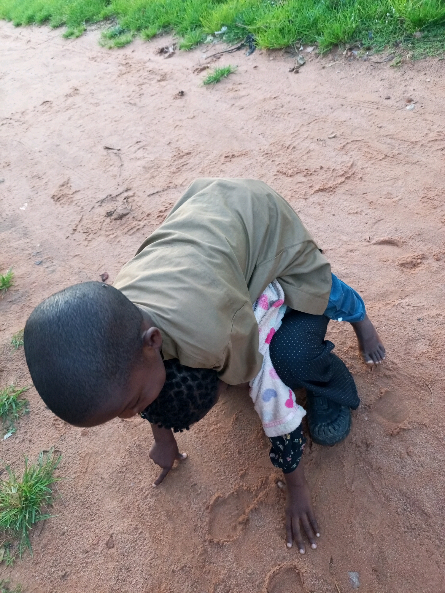 A child is on all fours in the dirt. They are wearing a green shirt and blue pants. A blanket is draped over their back. The child is reaching out with their right hand. The ground is made of reddish dirt with some patches of green grass. The image is taken from a high angle looking down at the child. The child is located in an outdoor, unpaved area in the city of Tempure, Nigeria.