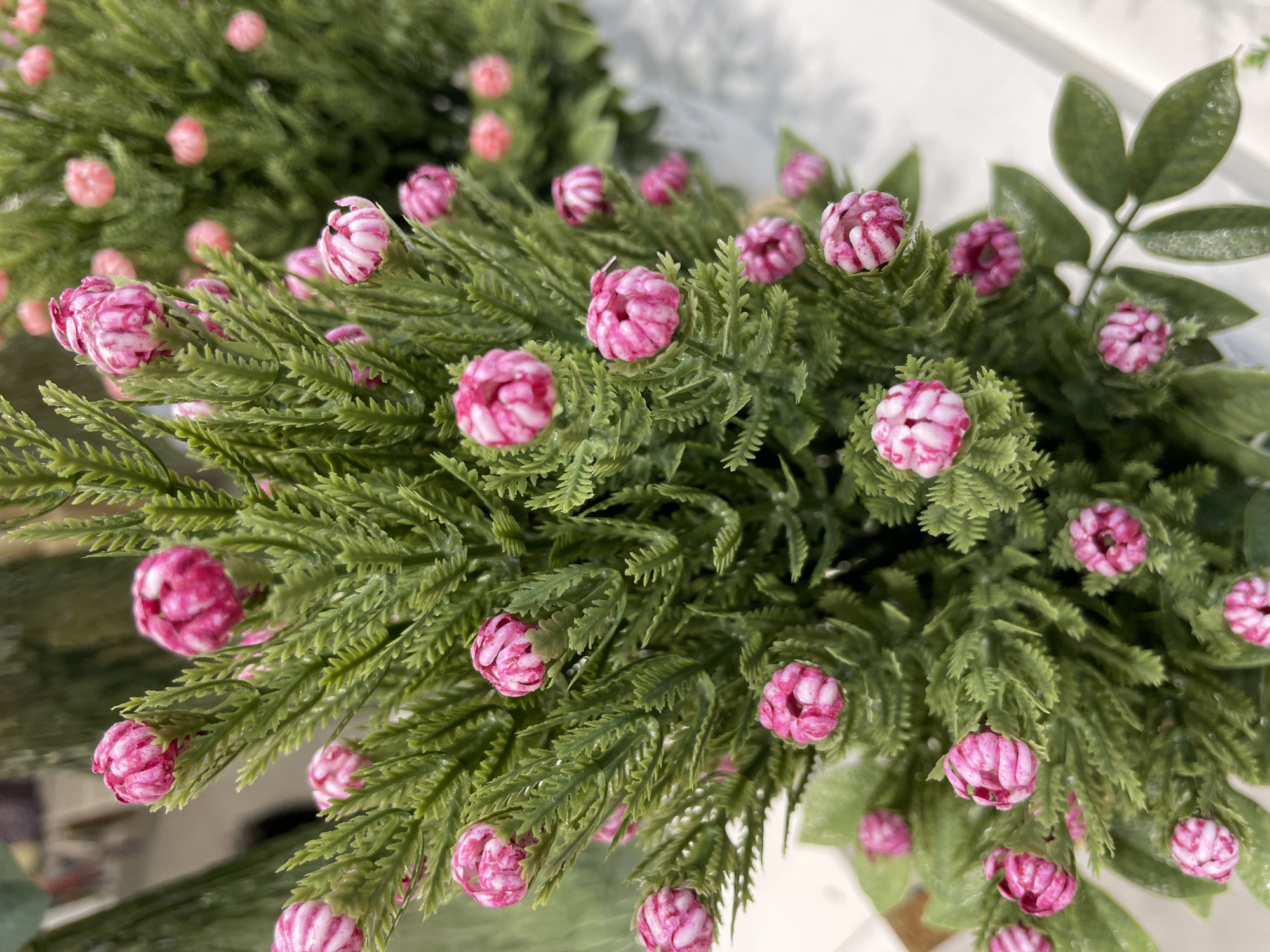 The image shows a close-up of a decorative plant with small, pink, bud-like flowers and green, needle-like leaves. The plant appears to be artificial and is arranged in a way that suggests it is part of a floral display or arrangement. There are other similar plants visible in the background, also out of focus. The lighting is bright and even, suggesting an indoor setting. There is no indication of the city of Perm, Russia, in the image itself.