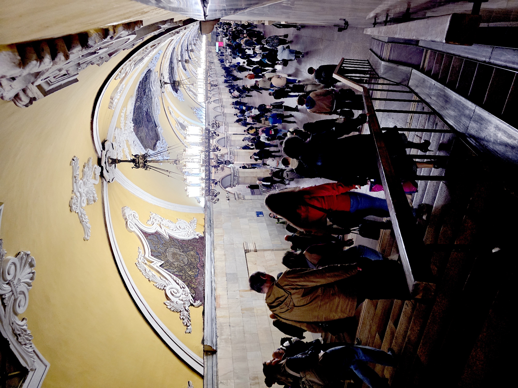 Here is a detailed description of the image:
The image shows a crowded scene in what appears to be a Moscow metro station, with an emphasis on architectural details and a large number of people. The perspective is somewhat unusual, looking down the length of the station and across to the stairs.
**Environment:**
- The station is characterized by its ornate, classical architecture.
- The walls are yellow with white embellishments, including decorative moldings and patterns. There are also mosaic designs on the walls.
- The station hall is well-lit with grand chandeliers.
**People:**
- A large crowd of people is present in the station hall.
- The crowd is densely packed, suggesting a busy time of day.
- People are dressed in typical casual wear.
**Objects:**
- Chandeliers hanging from the ceiling provide illumination.
- A staircase on the right side of the image descends.
- Escalators are visible beyond the staircase.
**Location:**
Given the architectural style, color scheme, and the presence of crowds, the image is most likely a platform in one of the Moscow Metro stations renowned for their opulent, Stalinist-era design, but it is not possible to provide the exact location of the station within Moscow.