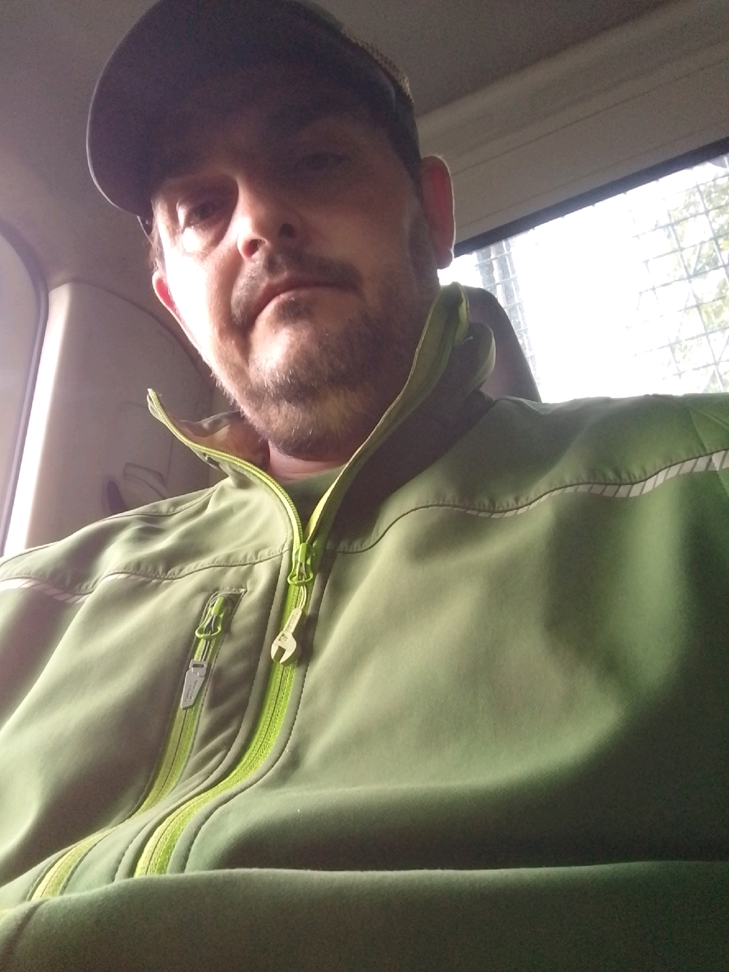 A man is seated inside a vehicle, seen from the chest up. He is wearing a green jacket with a lime green zipper. The jacket has reflective stripes near the shoulder. He is also wearing a dark baseball cap. The man has short, stubble-like facial hair and is looking directly at the camera. Behind him, a window shows a blurred, light-colored background, possibly indicating it is daytime. The interior of the vehicle appears to be a muted gray color. The image's lighting suggests it was taken indoors or in a shaded area.
