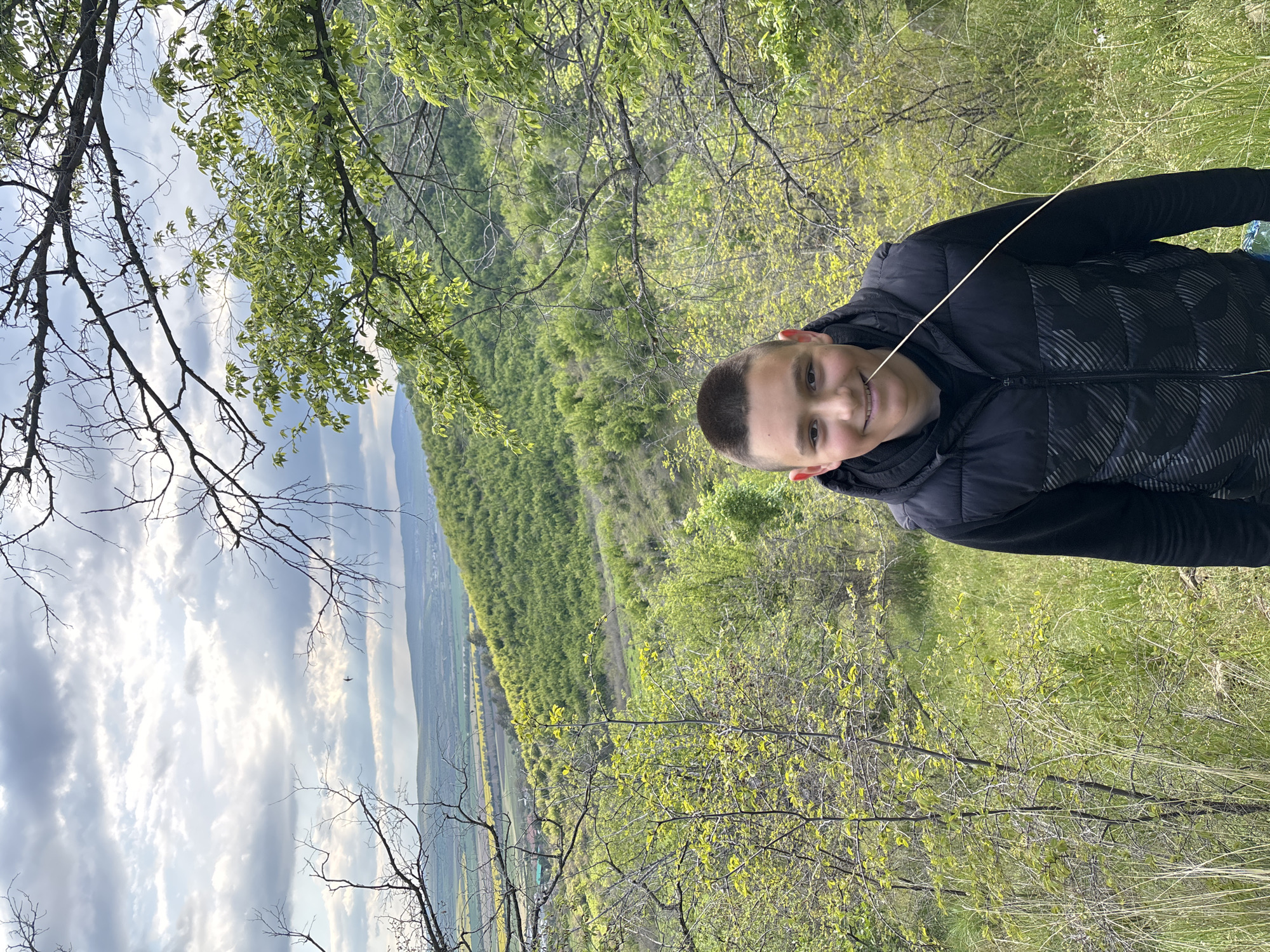 A medium shot depicts a boy standing outside in a hilly, rural area. He is smiling, wearing a dark, quilted vest and dark long-sleeved shirt. A blade of grass extends from one side of his face.
The backdrop is a sweeping landscape featuring a valley, partially visible through the trees. Dense, verdant foliage covers the hills. In the distance, a sliver of the sky is visible with scattered clouds, and traces of civilization, possibly part of the city of Goritsa, Bulgaria, can be seen in the valley. Branches of trees are visible, particularly in the foreground. The photo appears to have been taken from a high vantage point.