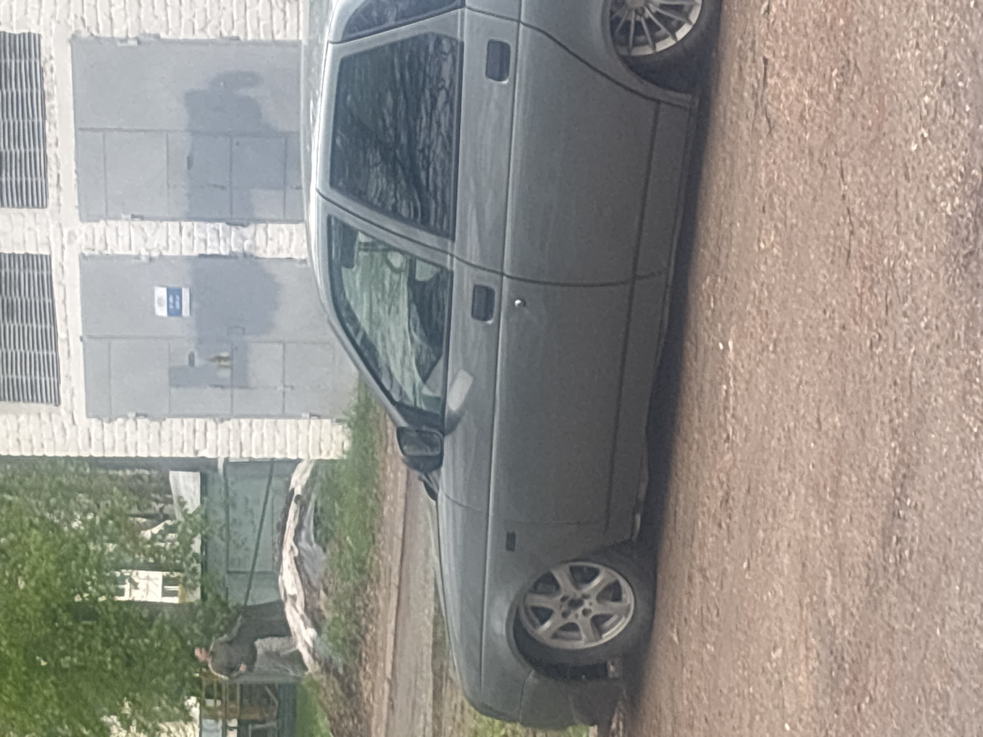 A grey four-door sedan is parked on a worn, dirt road. The car's wheels are silver, and the body appears to be in fair condition. To the left, a white brick building with grey metal doors is visible, along with some sparse greenery. In the background, a person wearing dark clothing can be seen standing near another structure. The environment suggests an industrial or semi-rural area. This could be a location in the city of Chaikovsky, Russia, although specific street information cannot be determined from this image.