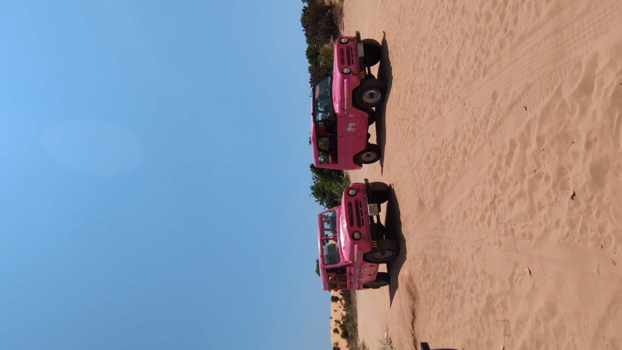 Two pink off-road vehicles are parked on a sandy terrain in Phuoc Dinh, Vietnam. The sky is clear and blue. Tire tracks are visible in the sand. Trees and vegetation appear in the background.
