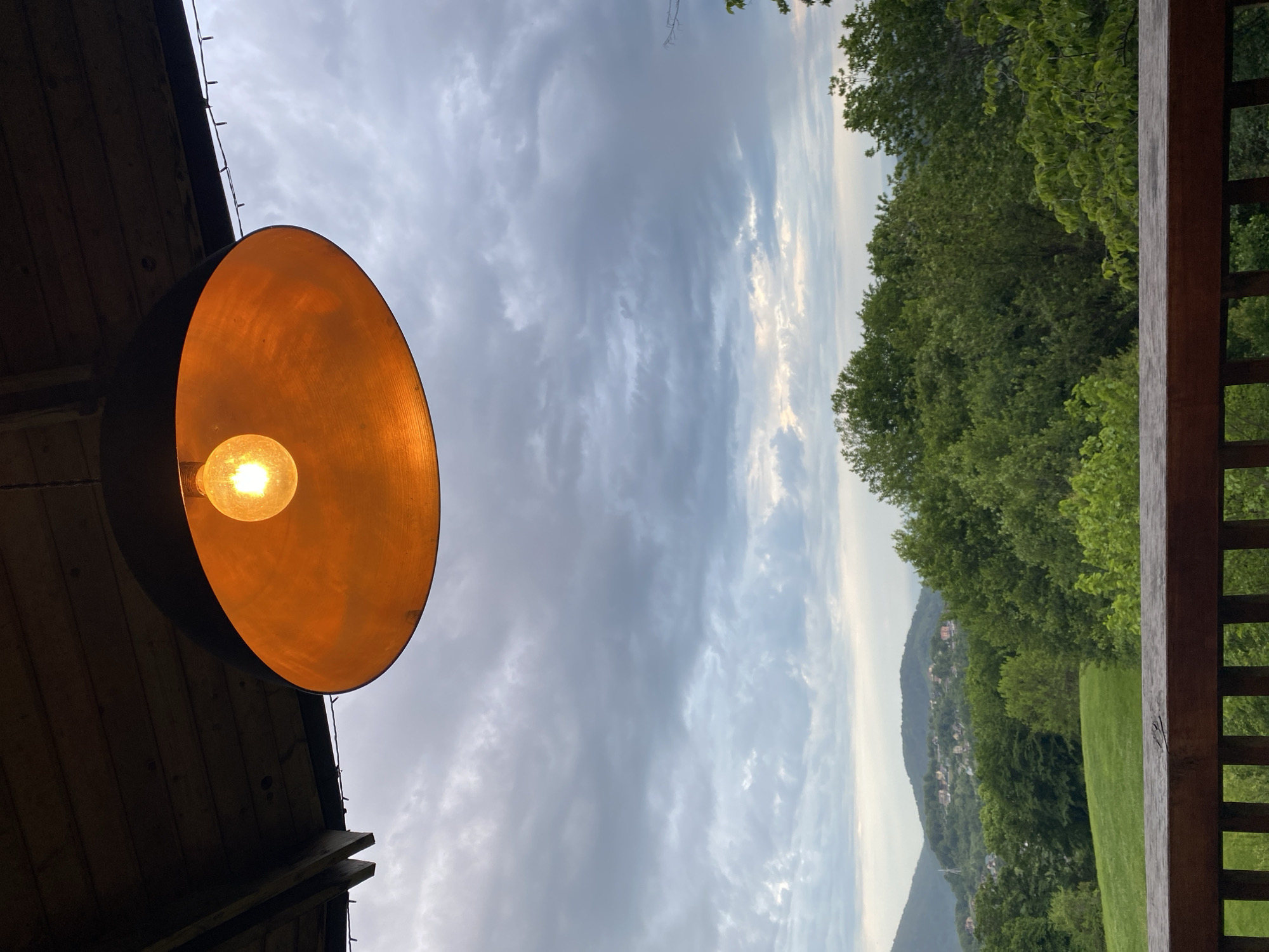 The image shows an outdoor scene under a cloudy sky, likely taken from a porch or deck. A large, round, orange-colored light fixture hangs prominently on the left side, illuminating the area. In the background, there is a green landscape with trees, grass, and hills. In the distant background, there are some buildings, likely part of a town or city. The scene appears to be in a rural or suburban area, not necessarily limited to Progres, Russia. The image does not depict any people or specific actions.