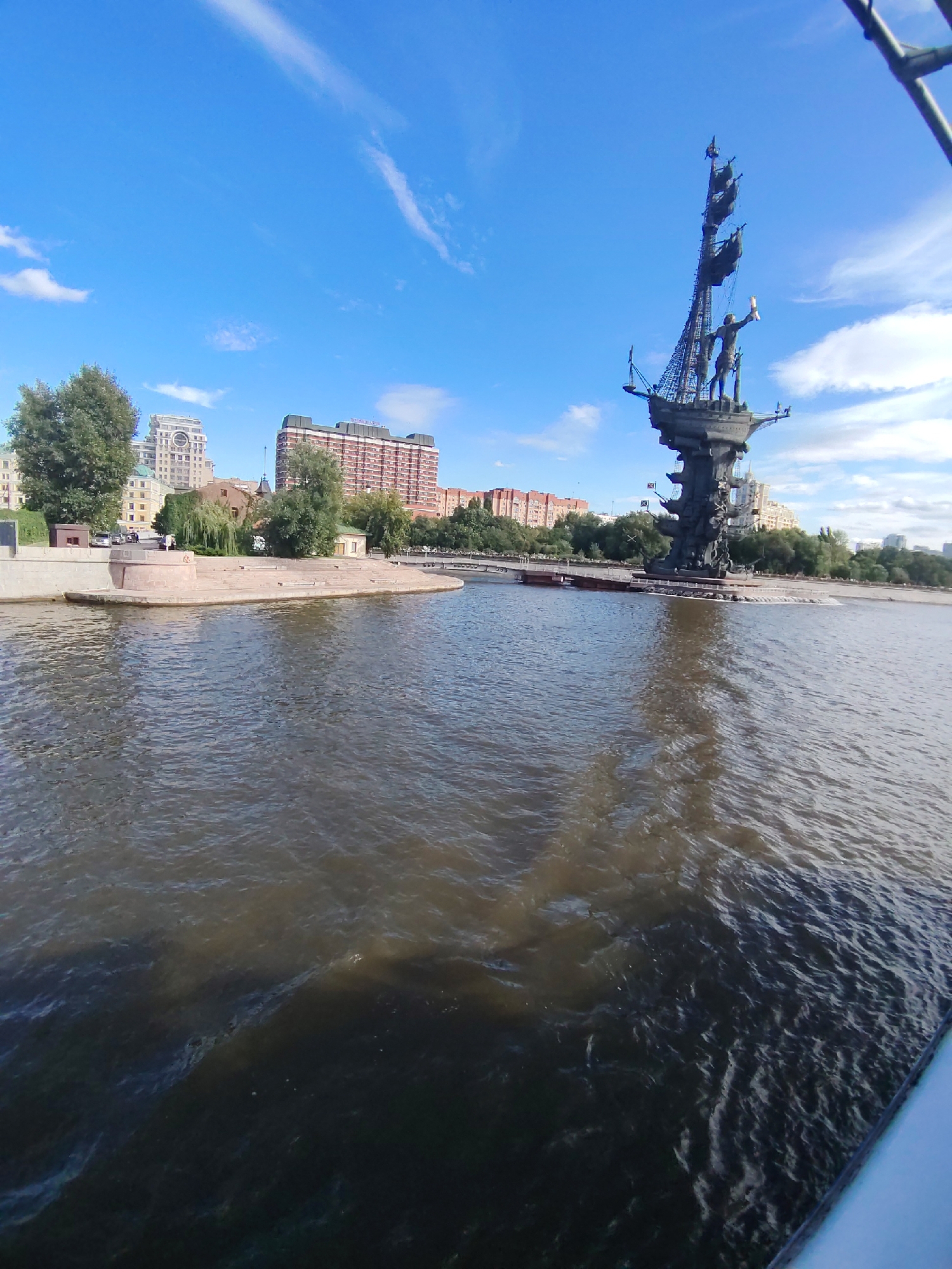 A monumental statue of Peter the Great on a ship is situated on the bank of a wide river, with city buildings and trees in the background under a clear blue sky with scattered clouds. The statue is dark-colored and depicts Peter the Great standing on the deck of a ship, holding a scroll or a sextant. The ship itself is detailed with masts and sails. The river in the foreground is a murky brown, reflecting the sky and the statue. On the riverbank, a stone embankment and a circular paved area are visible. Behind the embankment, several trees and a row of multi-story buildings are present. The buildings vary in architectural style, with some appearing more modern. The overall scene suggests a sunny day in a city with significant historical or artistic landmarks along a waterway.