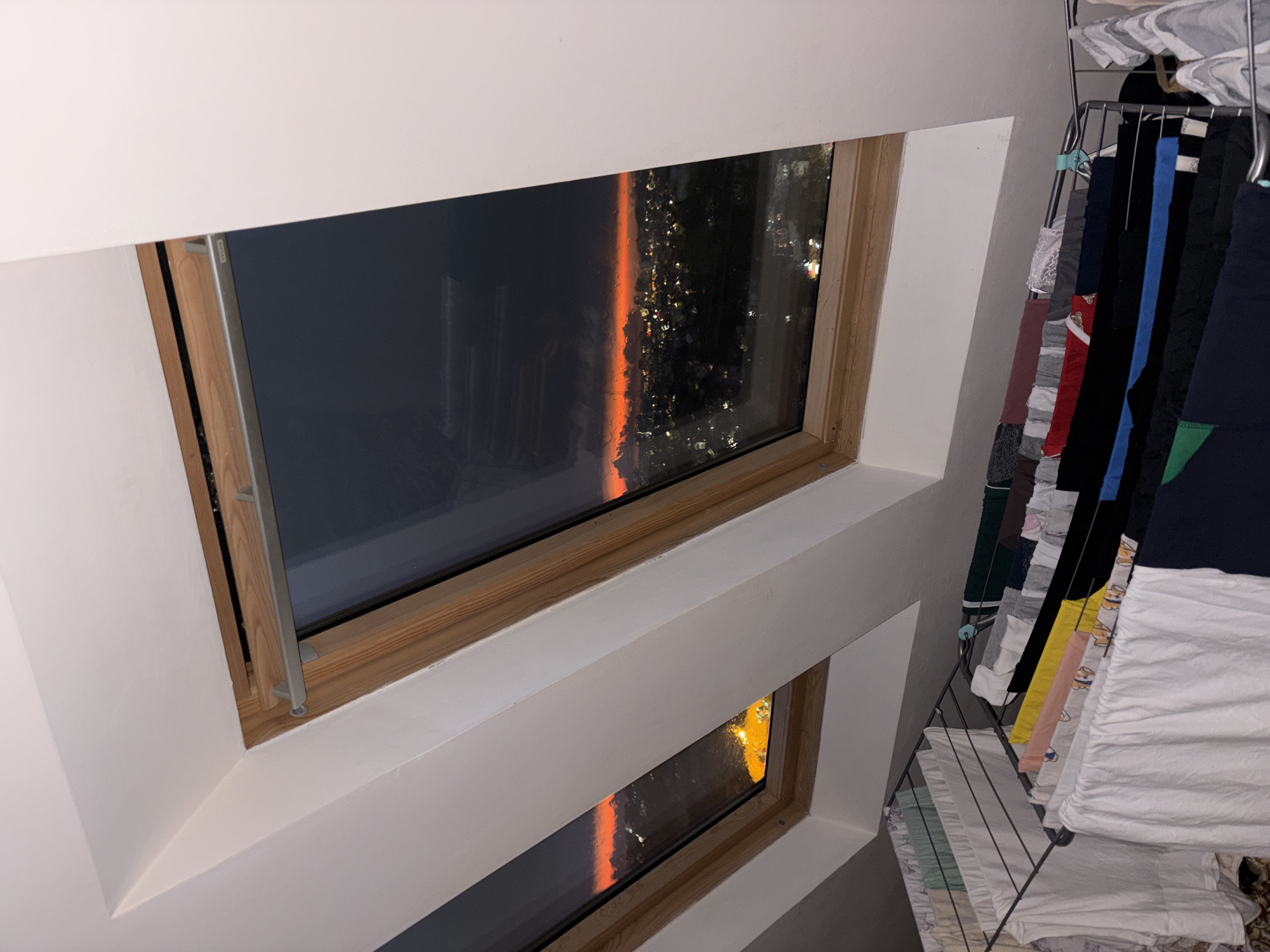 The image depicts a room with two rectangular windows, one above the other, built into a white wall. The window frames are light wood. Visible through the glass is a nighttime cityscape with bright orange reflections on the window, suggesting the scene is at sunset. A rack holding clothes is positioned to the right of the windows, near the wall. The clothes hanging on the rack vary in color and type.