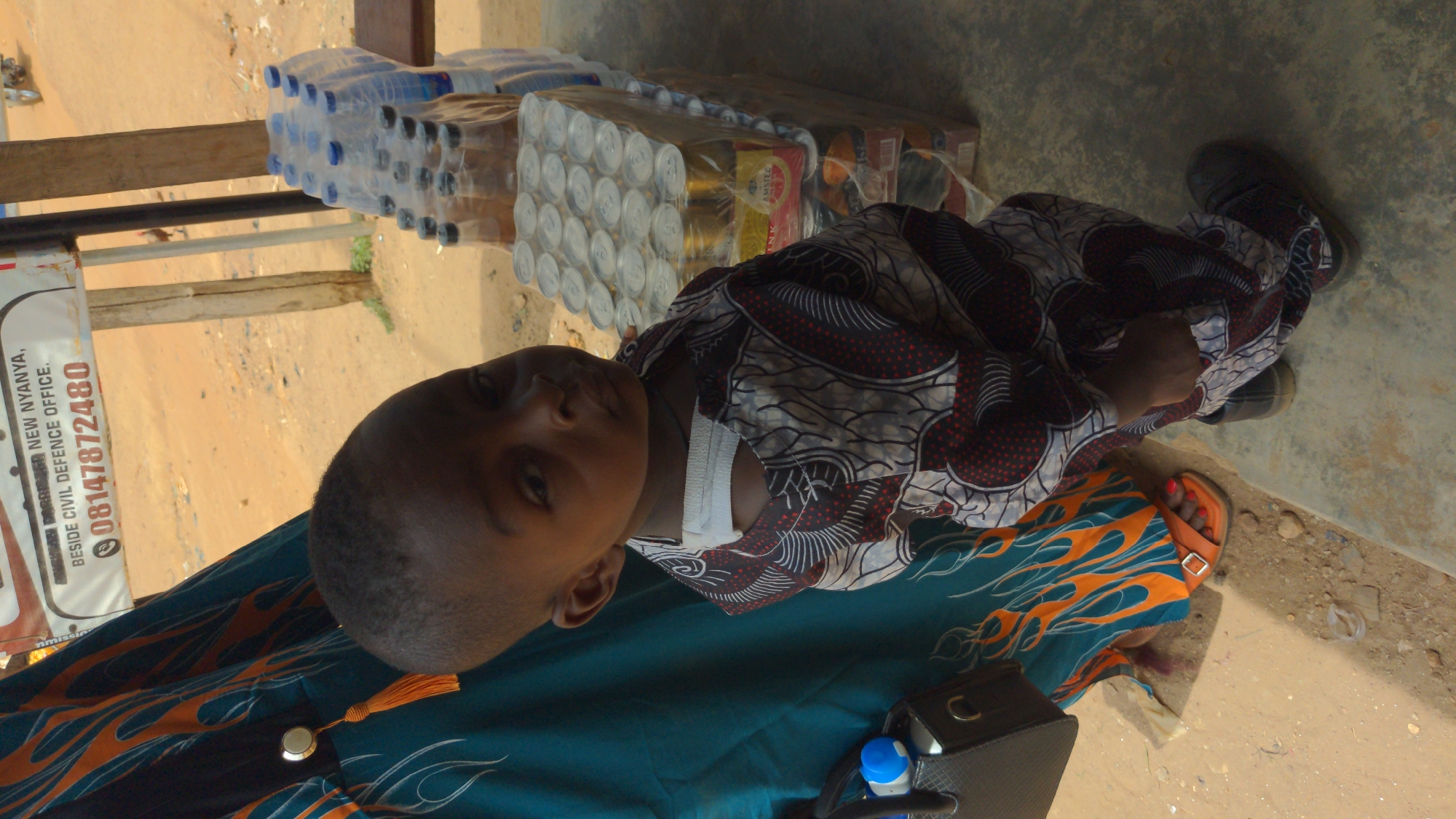 The image shows a child lying on an adult's lap. The child is wearing a patterned shirt. The adult is wearing a green and orange patterned outfit, orange sandals, and red nail polish on their toes. There are drinks piled up on a table near the child's head. A sign reading "BESIDE CIVIL DEFENCE OFFICE" and a phone number starting with "08147872480" is visible in the background. Based on the text on the sign, this may be near a Civil Defence office in Ado, Nigeria.