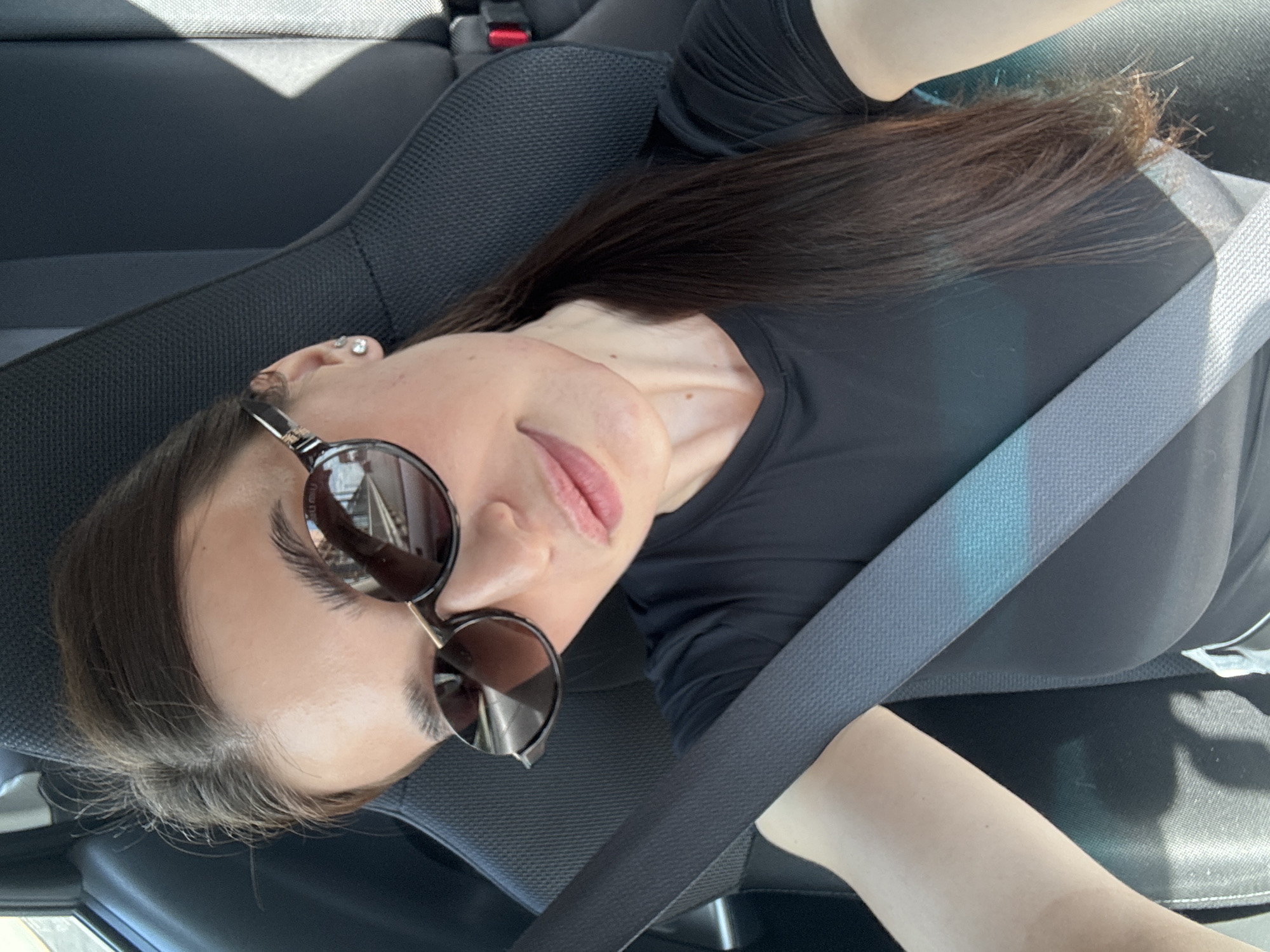 The image depicts a lone female individual taking a selfie inside a vehicle, in what is identified as the city of Chaikovsky, Russia. The subject, positioned in the front passenger seat, has dark brown hair styled in a low ponytail and is wearing oval-shaped sunglasses with dark lenses and frames accented with gold-tone hardware. A small, clear stud earring is visible in her right ear. She is wearing a black short-sleeved V-neck top and a gray fabric seatbelt diagonally across her torso and lap. Her lips are slightly parted with a neutral expression. The car interior features dark gray or black textured fabric seats. Strong daylight is evident, with sunlight casting a distinct diagonal shadow across the car's interior, particularly on the passenger seat and the seatbelt. Reflections in the sunglasses reveal an outdoor scene with vague structural elements. A red component, possibly a seatbelt release button, is partially visible on the far right edge of the frame, behind her.