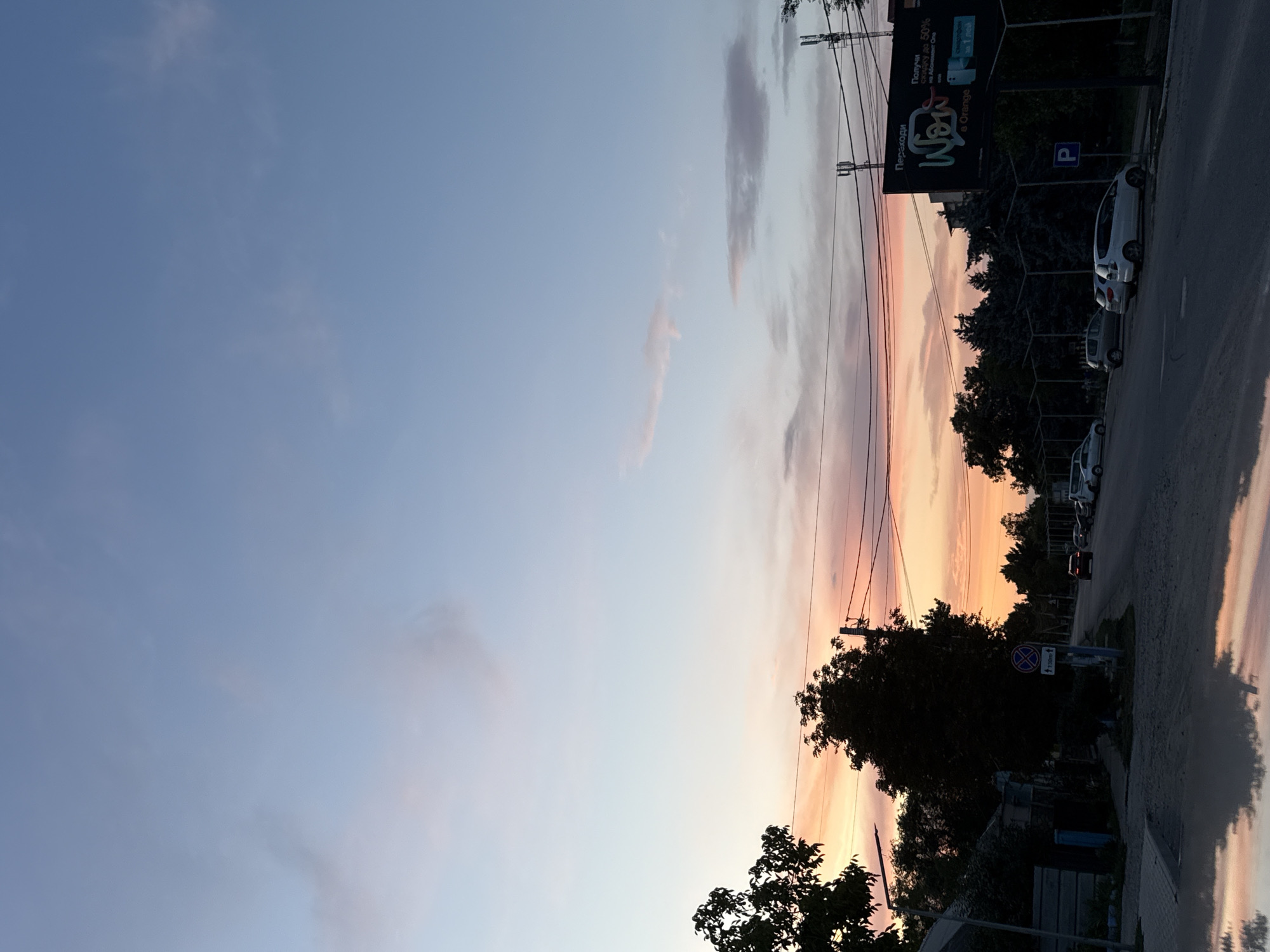 This image depicts a street scene at dusk in Basarabeasca, Moldova. The sky is a gradient of soft blues and pinks, indicating the setting sun. Silhouetted trees line the sides of the road, with some appearing to be in the foreground on the left, and a denser line of trees further down the street and on the right. A streetlamp is visible on the left. Several cars are parked along the right side of the road, and a few are driving on the road. A large billboard is mounted on the right, displaying text in a foreign language and graphics, suggesting it's an advertisement. The road surface appears wet, possibly from recent rain, with reflections visible on the ground in the foreground, suggesting a puddle or accumulated water. Power lines are strung overhead, supported by poles. A circular traffic sign with a diagonal red line is visible on the left, and a blue parking sign is visible on the right. The overall impression is a quiet, residential street at the end of the day.