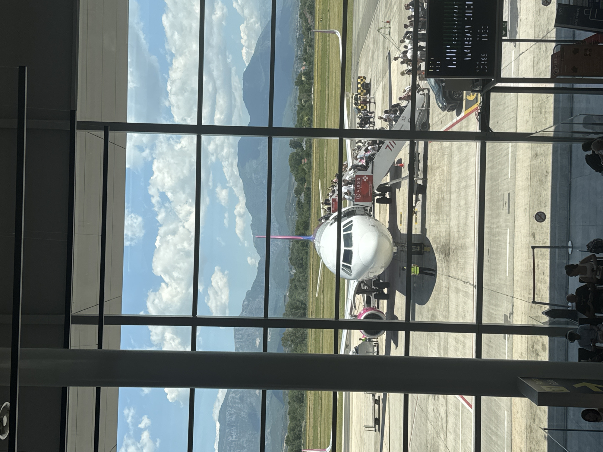 The image depicts an airport tarmac scene, viewed from behind a large window with vertical and horizontal mullions. In the foreground, a large airplane, identified by its pink and white livery as likely a Wizz Air aircraft, is parked on the tarmac. Boarding stairs are attached to the front of the plane, with numerous passengers visible ascending and descending. A ground crew member in a yellow vest stands near the airplane's engine.
In the background, a mountainous landscape with a clear blue sky dotted with white clouds stretches across the horizon. Green fields and trees are visible between the tarmac and the mountains.
The airport environment is evidenced by the presence of the aircraft, tarmac markings, baggage carts, a service vehicle, and other airport equipment. People are seen in various states of boarding and disembarking the plane, indicating an active airport operation.
The question mentions the city of Kruja, Albania. While the image itself does not provide direct visual cues to confirm this location, the presence of a Wizz Air aircraft suggests a European context. If this scene is indeed in Kruja, Albania, it would indicate the presence of an airport serving that region. The overall scene is one of travel and transit within an airport setting, with a backdrop of natural mountain scenery.