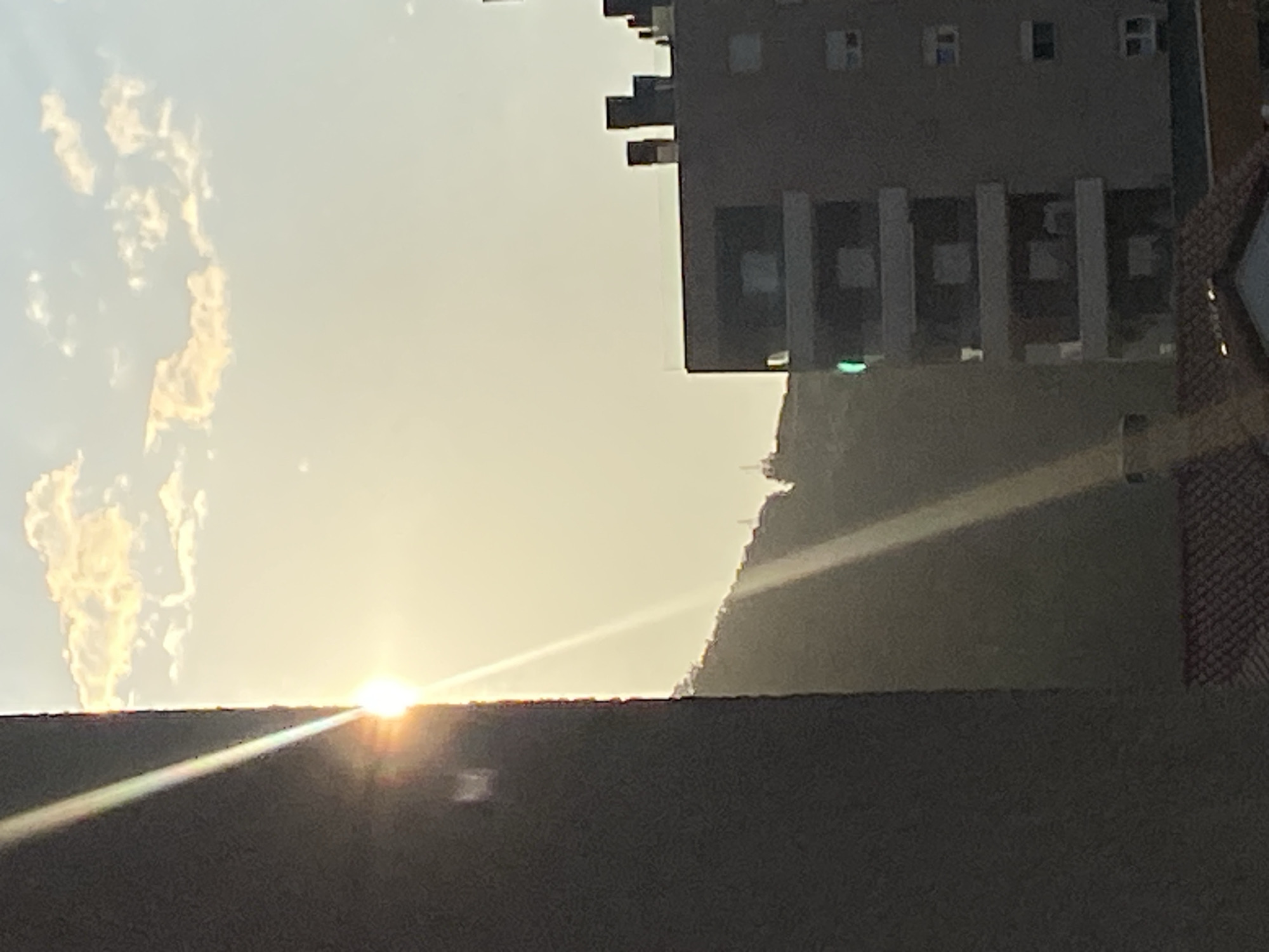 The image shows a cityscape at dawn or dusk, possibly in Budva, Montenegro. A modern multi-story building with prominent columns stands against a backdrop of a sloping hill or mountain partially covered in vegetation. The sun, low on the horizon, shines brightly, creating a strong glare and light rays. The sky is hazy, with some cloud formations visible. A dark, out-of-focus structure, possibly a rooftop or wall, dominates the foreground. The orientation of the photograph is unusual, potentially rotated, affecting the perception of spatial relationships.