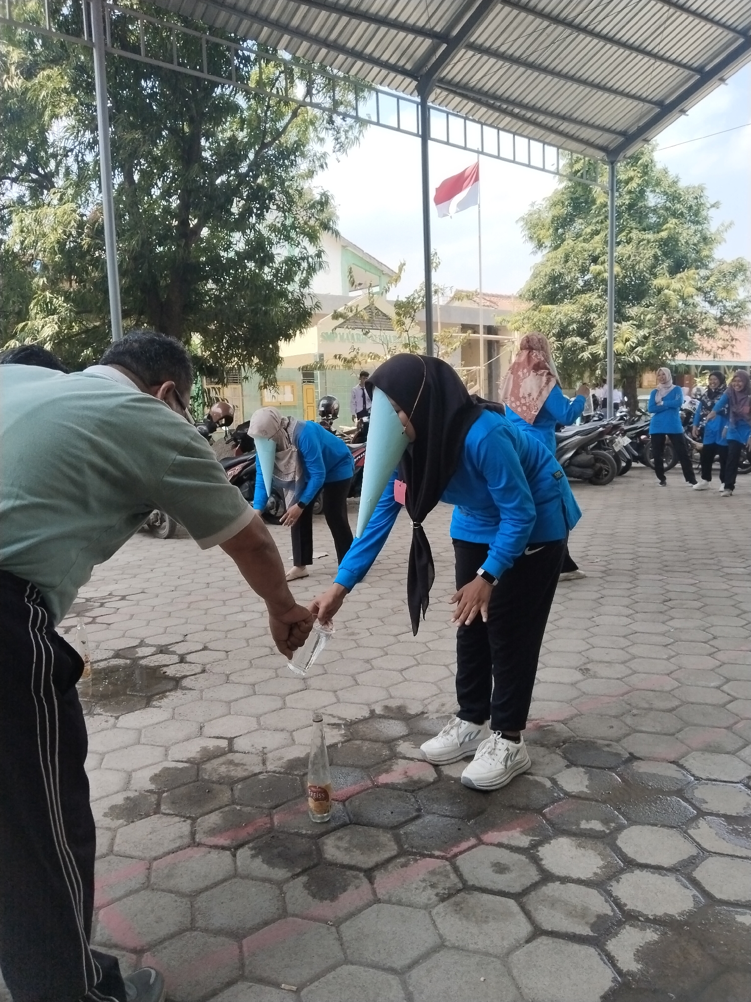 In the city of Pakulaut, Indonesia, under a covered outdoor area with a metal roof supported by metal poles, an event is taking place on a hexagonal paver stone ground, wet in places. In the foreground, a man wearing a light green t-shirt and black pants with white stripes is holding a clear plastic bottle in his right hand and leaning over to pour its contents into an empty glass bottle with a label that reads "FREISS," positioned on the ground.
To his right, a woman wearing a blue long-sleeved shirt, black pants, white sneakers, and a light blue conical head covering with black tassels is bent over, reaching towards the ground. Behind her, another woman in a similar blue shirt and head covering is standing, and further back, several other women in blue shirts and hijabs are visible, some standing and some with their backs to the camera. The Indonesian flag is visible flying on a flagpole in the background, along with trees and buildings, suggesting a school or public facility. Motorcycles are parked to the right of the scene. The overall atmosphere appears to be a recreational activity or competition.