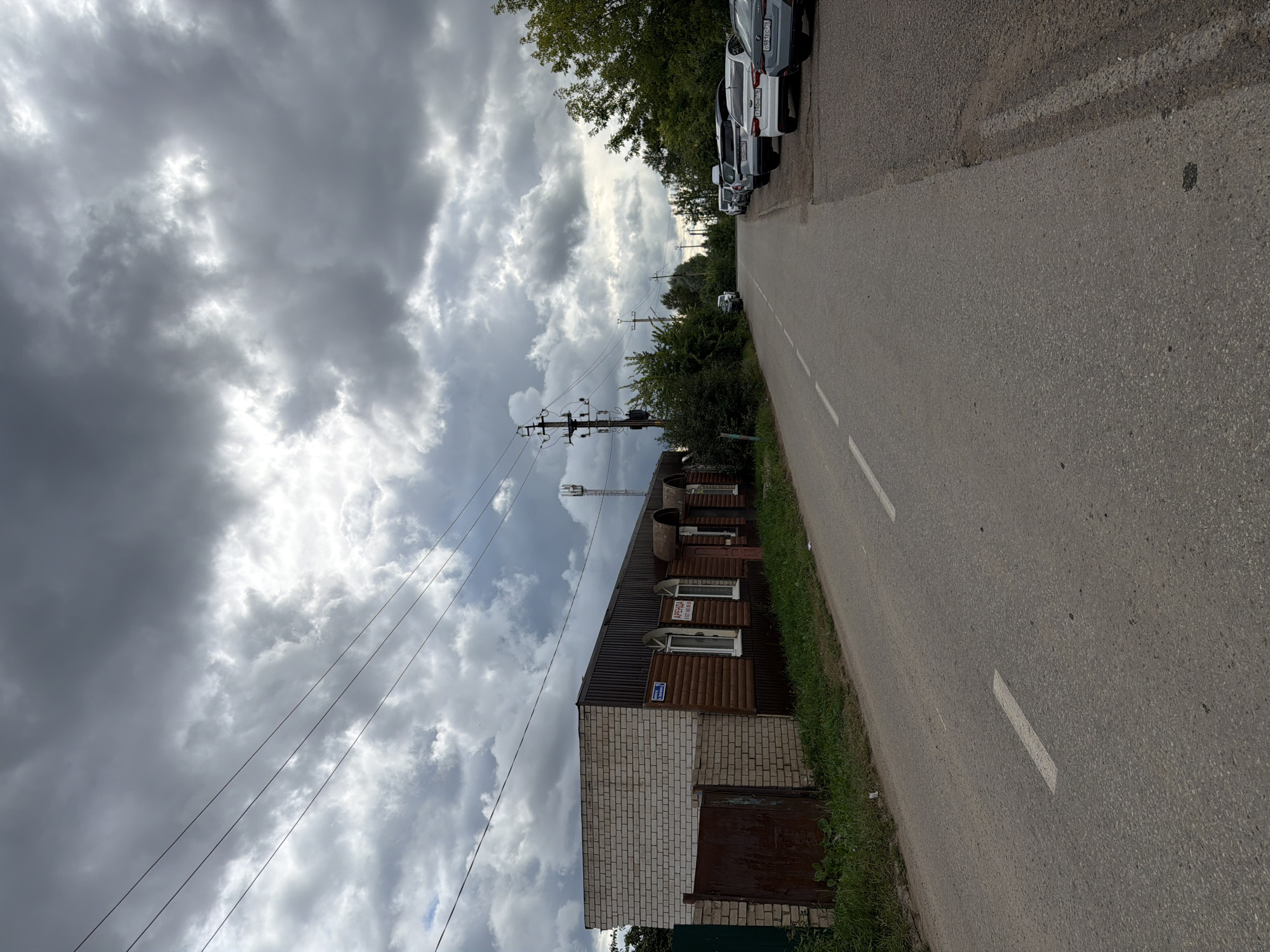 The image depicts a cloudy day in Yelabuga, Russia. A paved road runs from the foreground into the distance. On the left side of the road, there is a low-rise building with a white brick front and a wooden siding facade, featuring arched windows and a rusty metal gate. On the right side of the road, several cars are parked along the curb. Trees and vegetation line both sides of the road. A telecommunications tower is visible in the background. The sky is filled with dark, dramatic clouds.