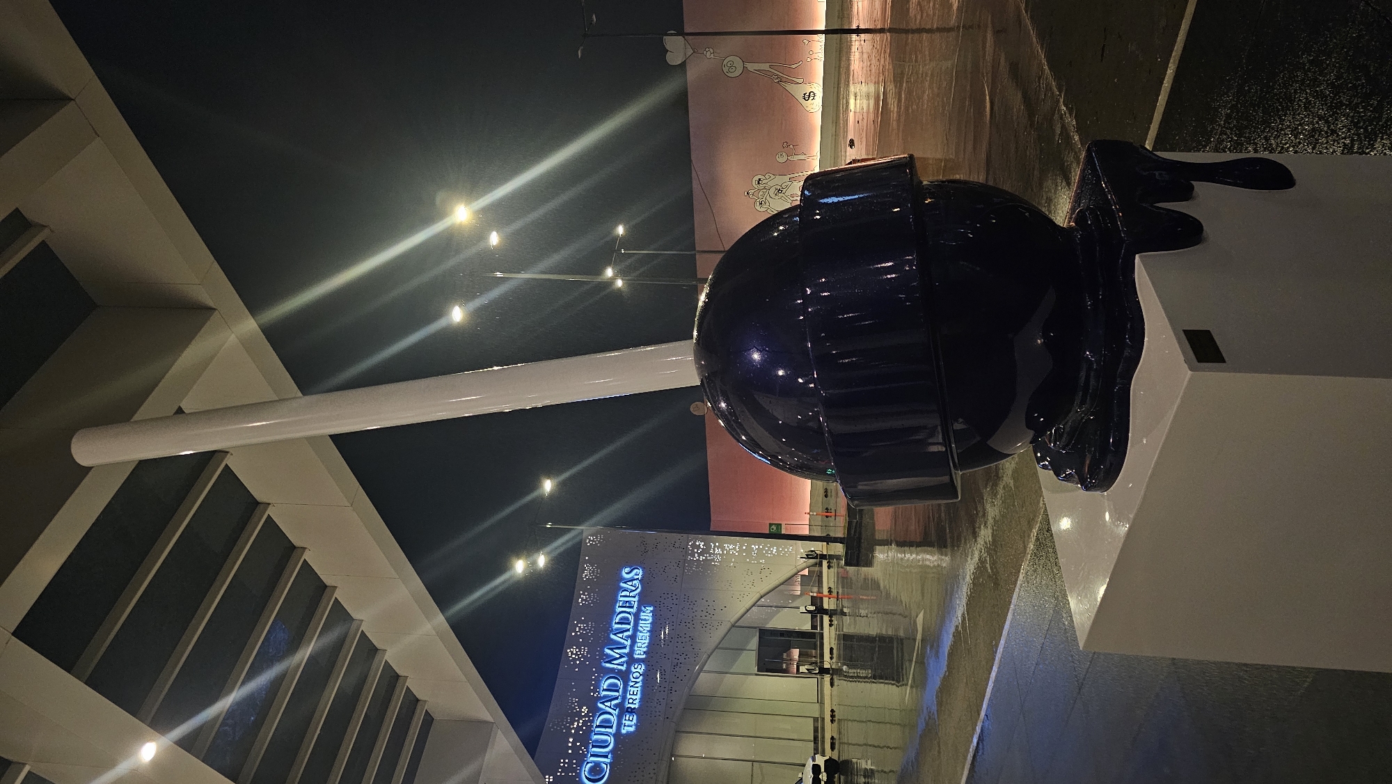 The image depicts a large, dark, shiny lollipop sculpture on a white pedestal. The lollipop's 'melting' effect suggests an artistic design. The backdrop includes a building with the words "Ciudad Maderas TE RENOS PREMIUM" illuminated in blue. Overhead lighting suggests it is nighttime. In the background, a wall with cartoon-like graffiti is visible. The ground appears wet, possibly due to rain. The image is taken at an angle. The location is Ciudad Maderas in Puebla, Mexico. No people are visibly present.