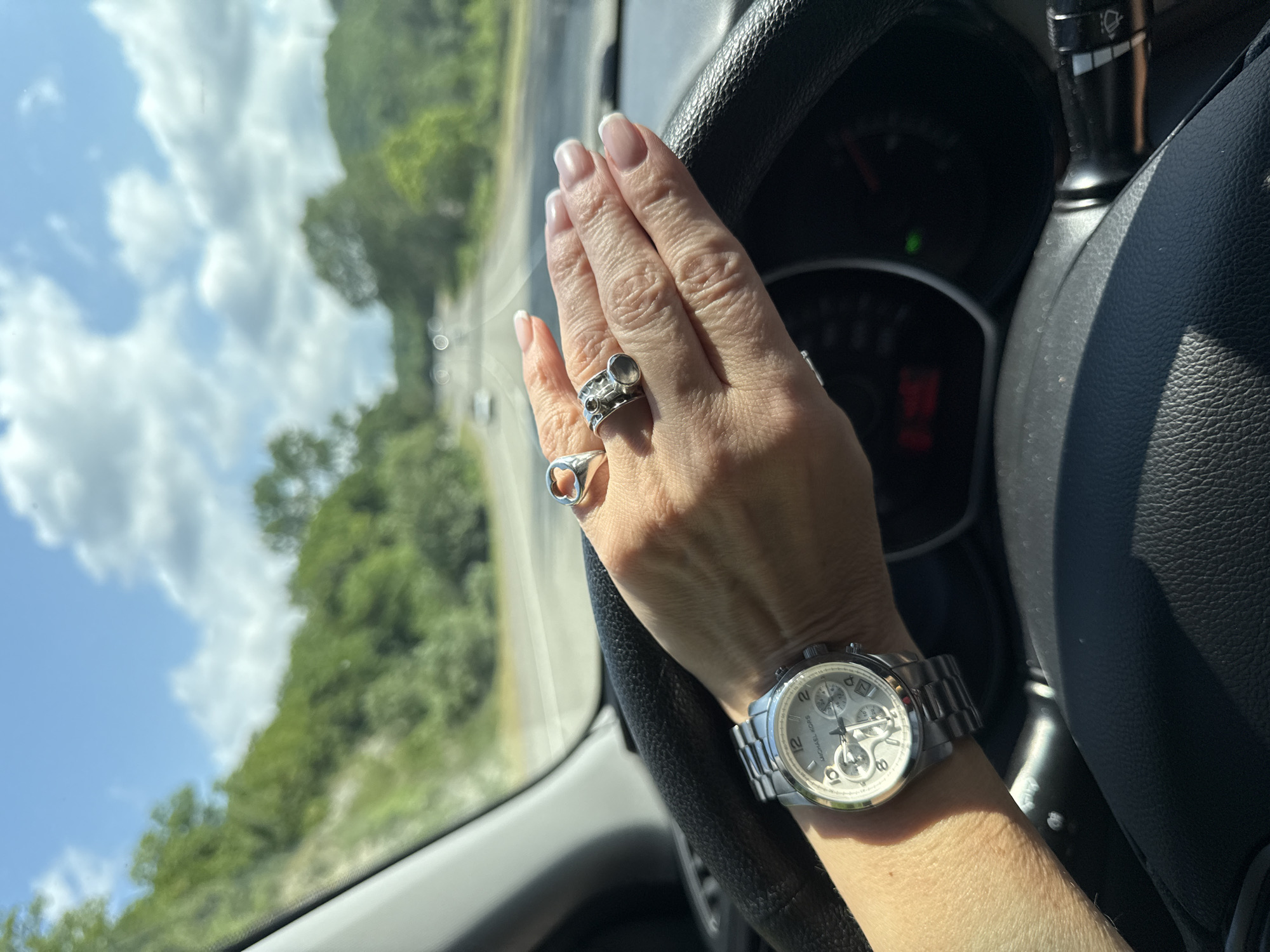 The image shows a close-up of a person's hands on a car's steering wheel. The person's right hand is resting on the steering wheel, adorned with several rings and a silver watch with a white face. The watch has subdials and the brand name "MICHAEL KORS" is visible. The person's fingernails are manicured with a French tip. The steering wheel itself is black and textured.
The background, visible through the car's window, suggests a scenic drive. There are lush green trees and a bright blue sky with fluffy white clouds, indicating fair weather. The road is curving, and there are hints of other vehicles in the distance. The lighting suggests it is daytime, likely late morning or early afternoon, given the bright sunlight.
The scene appears to be a person driving a car, possibly on a road trip or a leisurely drive through a natural landscape. There are no overt emotional cues, but the hands appear relaxed on the steering wheel. The image is likely taken from inside the car, looking towards the driver and the road ahead. There is no visible text in the image other than the brand name on the watch. The location context provided is Novomikhailoskiy, Russia.