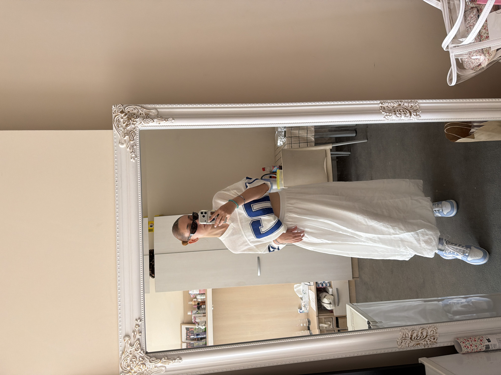 A person stands in front of a large, ornate, white-framed mirror, holding a phone and taking a picture of themselves. The person wears a white cropped athletic jersey with blue numbers, a long white skirt, and light blue sneakers. The background reflected in the mirror shows a room with light-colored walls, a white cabinet, shelving with various items, a white chair, and a gray floor. A partial view of a bag hangs near the top right corner of the mirror. The location is not discernible from the image.
