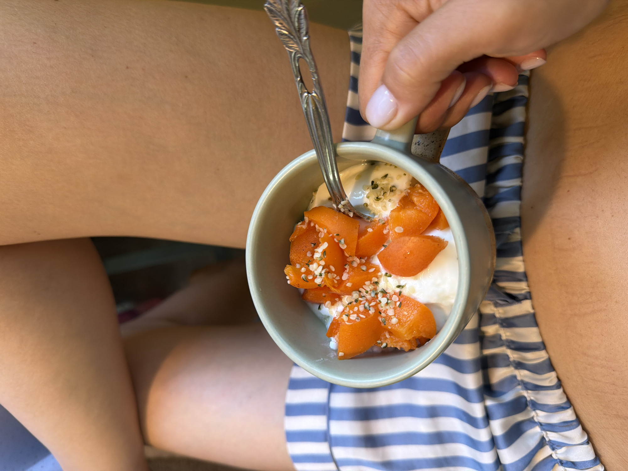 A close-up shot shows a person's thighs and a bowl of yogurt topped with sliced apricots and seeds. The person is wearing blue and white striped pajama bottoms. A spoon is resting in the bowl, and a hand with light pink nail polish is holding the bowl. The background is out of focus and appears to be an indoor setting. There is no identifiable information within the image to suggest a location in Pleven, Bulgaria.