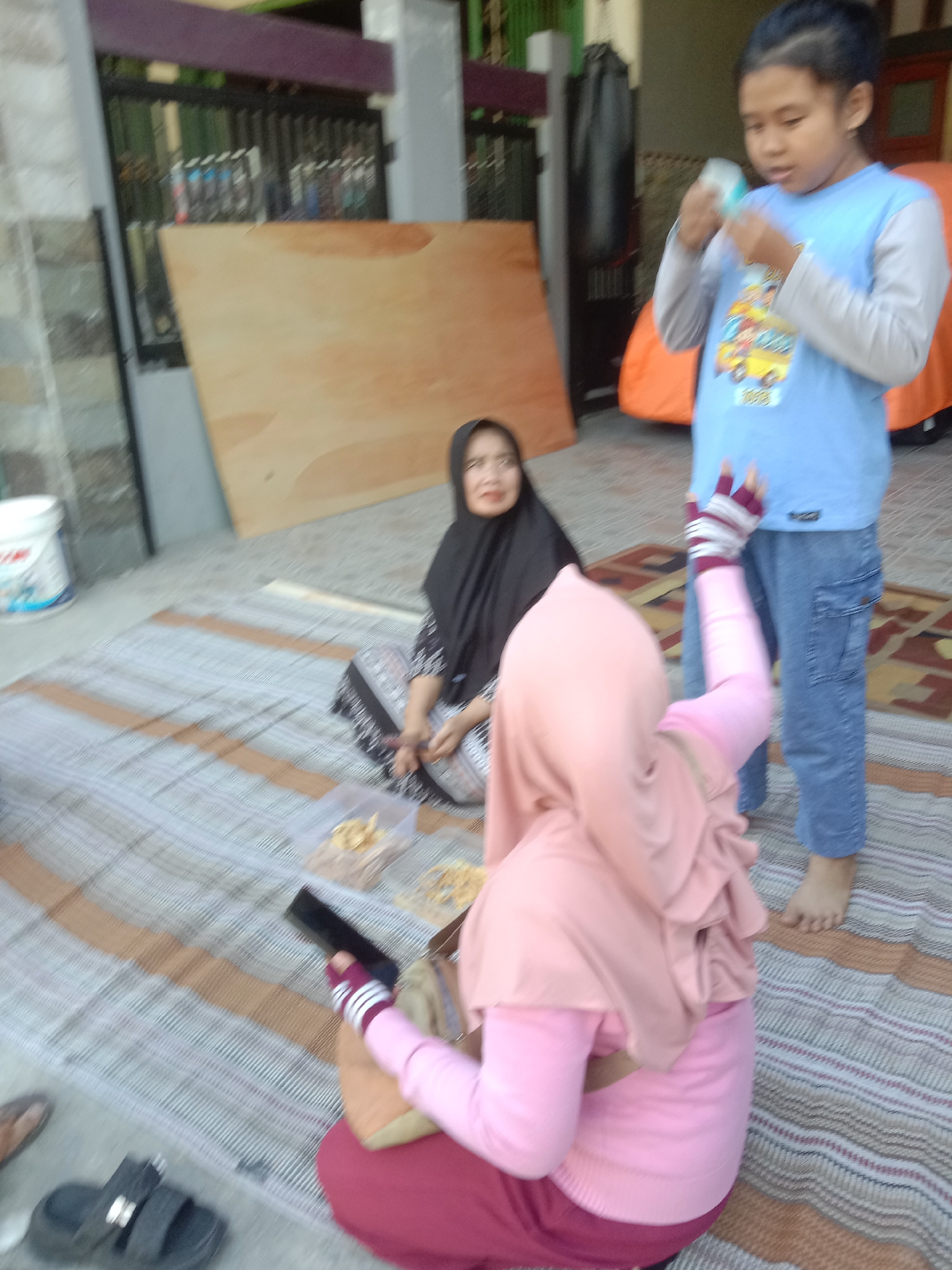 The image depicts a street scene in Tulungrejo, Indonesia, with an emphasis on a group of people engaged in an activity on the ground. In the foreground, a person wearing a pink hijab and pink long-sleeved shirt is seated, holding a dark object, possibly a phone. To their left, a person in a black hijab and patterned clothing sits on a mat, looking towards the viewer. To the right, a child in a blue long-sleeved shirt with a graphic print is standing and appears to be interacting with something in their hand. The ground is covered with mats, and several clear plastic containers filled with food items are visible. In the background, there are building structures, a wooden board, and an orange tent-like object. The overall lighting suggests an outdoor setting, possibly during the day.