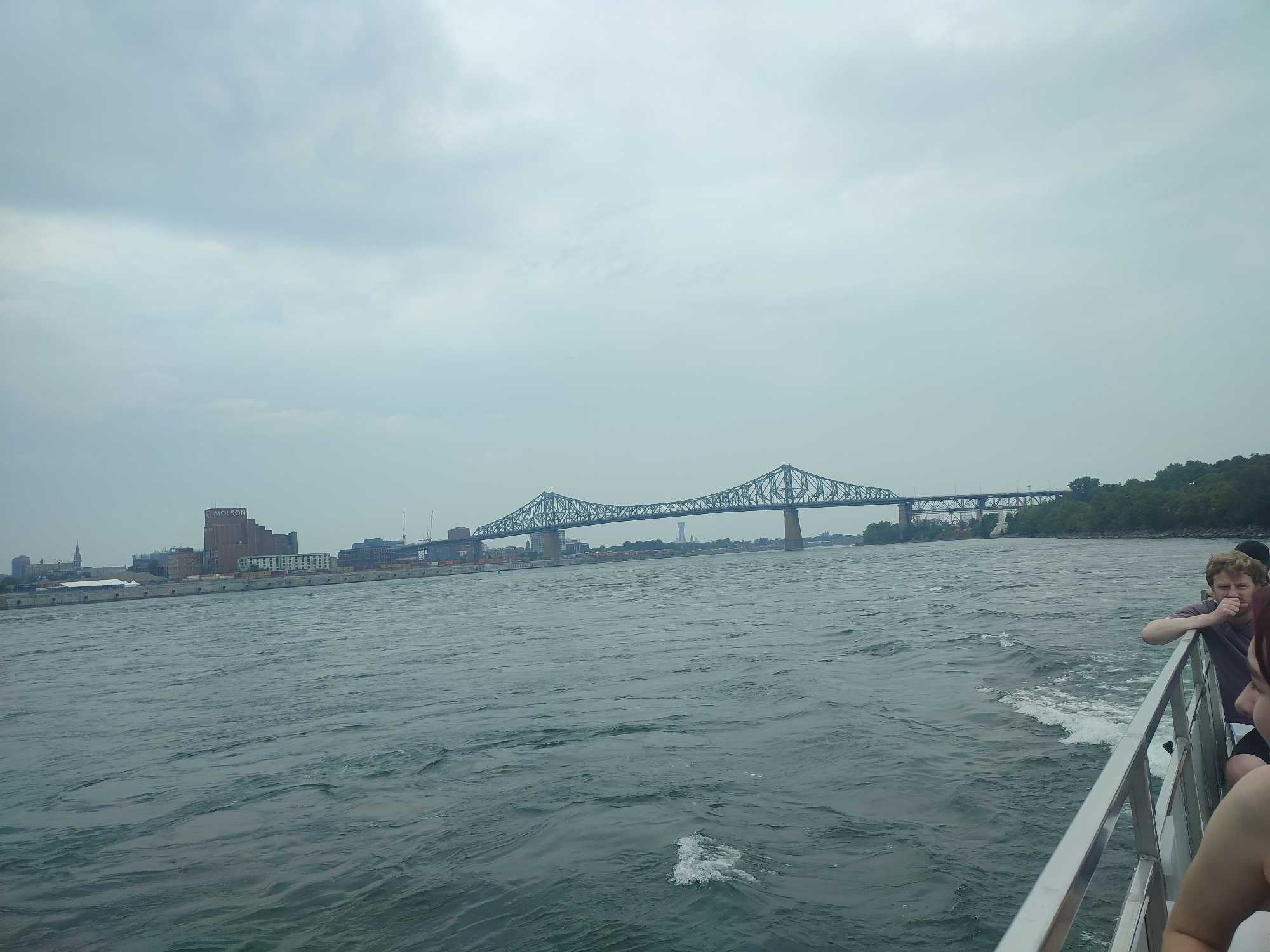 This is a photograph taken from a boat on the Saint Lawrence River in Montreal, Canada, on a cloudy day. In the background, the iconic Jacques Cartier Bridge spans the river. The city skyline is visible on the north shore, featuring industrial buildings, including a prominent "MOLSON" brewery building. On the right side of the frame, the railing of the boat is visible, along with a few individuals. One person, a male with reddish hair and a beard, leans on the railing, looking towards the camera with a hand near his mouth. Another person, partially visible, is seated to his right. The water is choppy with whitecaps, indicating movement of the boat. Trees and a green shoreline are visible on the far bank of the river, to the right of the bridge.