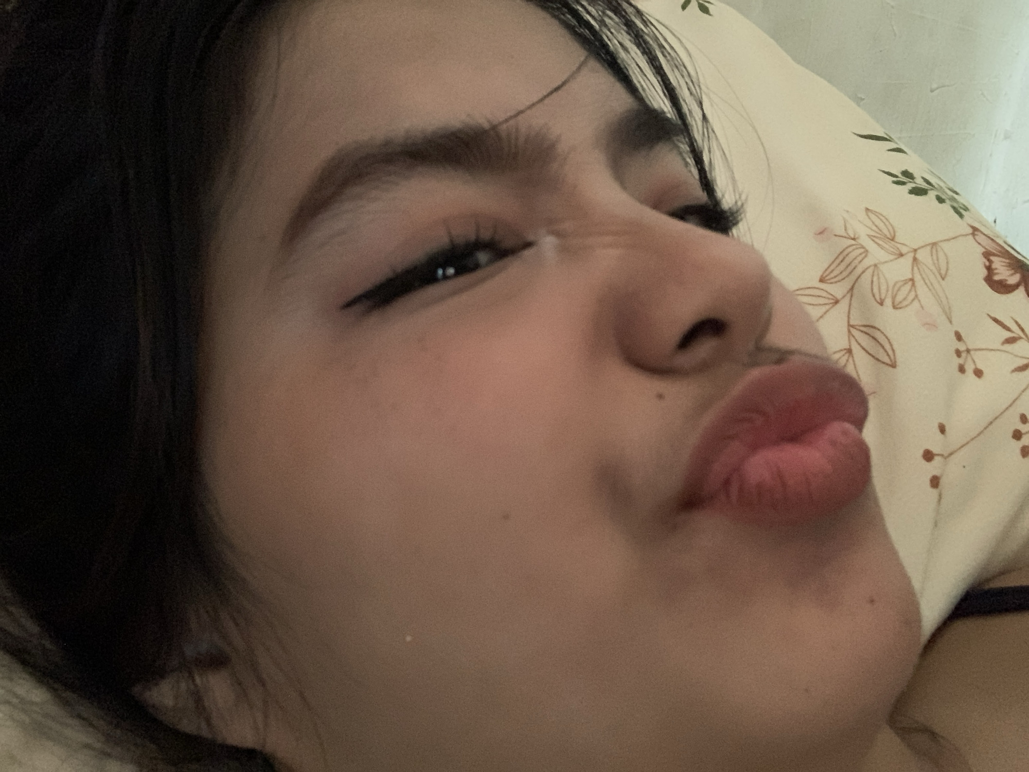 A close-up, diagonal shot captures the upper right side of a person's face, making a playful "kissing face" expression. The individual, likely female, has dark hair that frames the face and dark, well-defined eyebrows. One eye is visible, angled and partially closed due to the expression, with a noticeable winged eyeliner applied. The nose is slightly scrunched, and the lips are puckered outward, painted with a natural pinkish hue.
The skin tone appears warm, with a few small moles or beauty marks visible on the cheek and chin area. The background primarily consists of a light-colored pillow with a subtle floral and leafy pattern in shades of green and brown, suggesting an indoor setting, possibly a bedroom. A thin dark strap, possibly from clothing, is barely visible on the far right edge of the frame. The lighting is soft, creating a gentle ambiance. No text is visible in the image. The overall mood is intimate and lighthearted.
