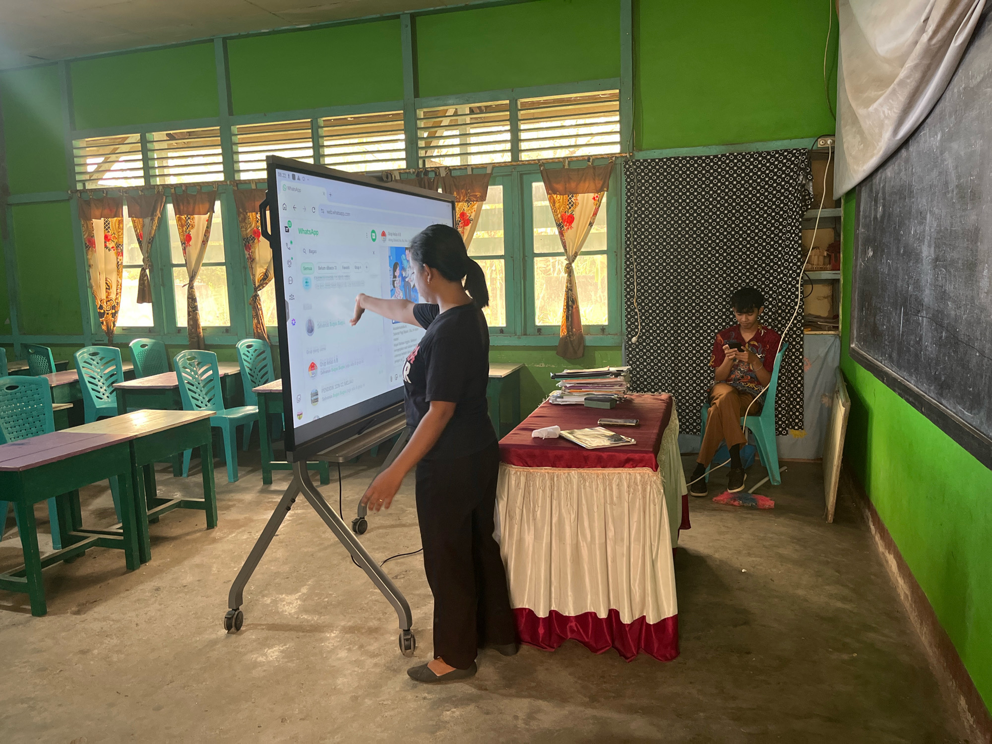 The image depicts a classroom setting in what appears to be a school in Meliau Hulu, Indonesia. A woman, dressed in a black t-shirt and dark pants, stands in front of a large, interactive display screen, pointing at it with her right hand. The screen displays a WhatsApp interface. The classroom is equipped with green desks and chairs, some of which are stacked. Curtains hang in the windows, and the walls are painted green. To the right of the woman, a young person, possibly a student, is seated in a blue chair and looking at a mobile phone. A table draped with a red and white cloth is positioned between the woman and the seated individual. A blackboard is visible on the far right wall. The flooring is concrete, and the overall environment suggests a functional, albeit somewhat dated, educational space.