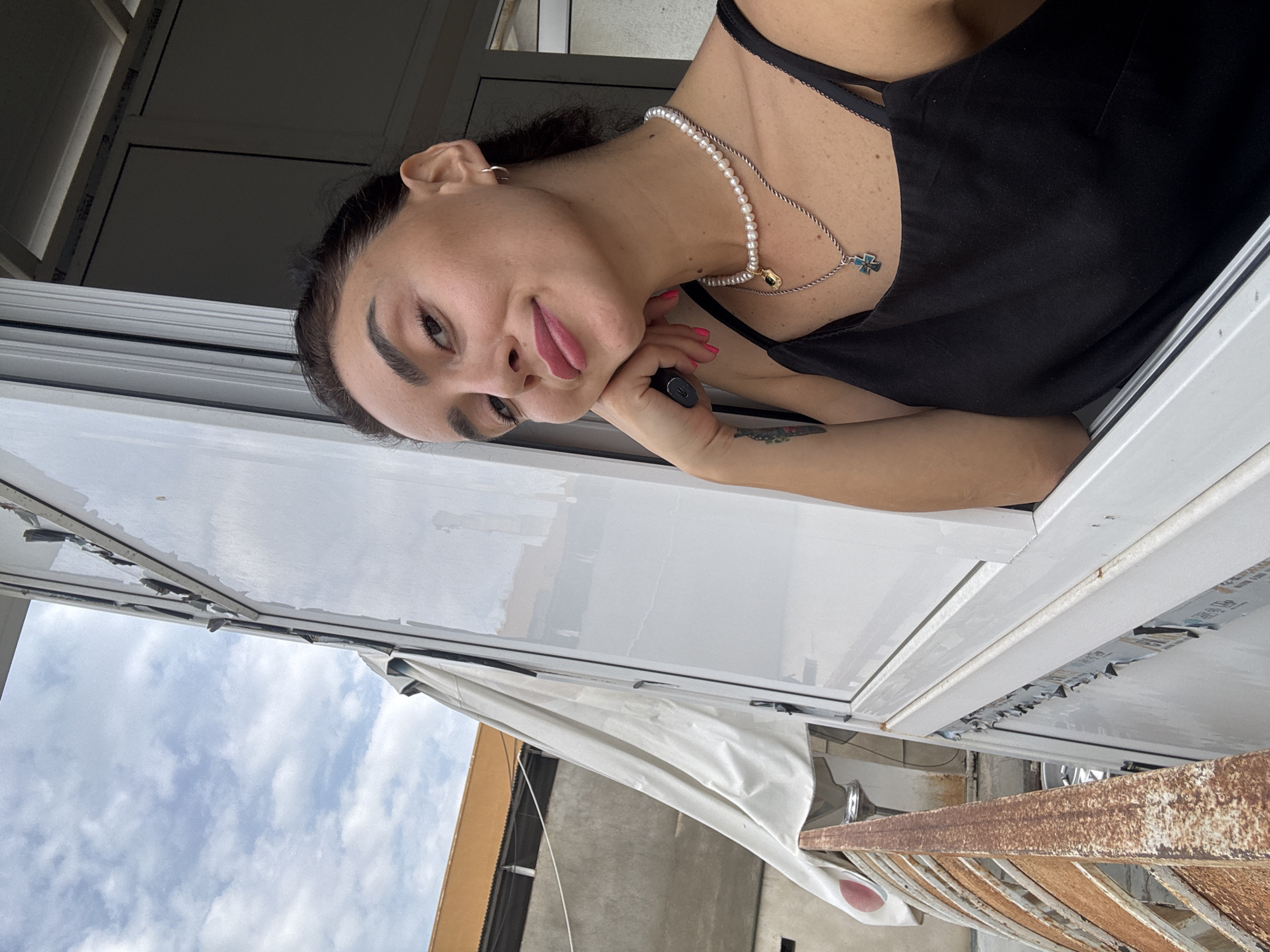 A woman is taking a selfie from an open window, leaning her head on her hand. She is wearing a black tank top, a pearl necklace, and a silver necklace with a cross pendant. The background shows a cloudy sky and buildings. The woman has dark hair pulled back, dark eyebrows, and is wearing pink lipstick. Her fingernails are painted pink. A rusted railing is visible to the left of the frame.