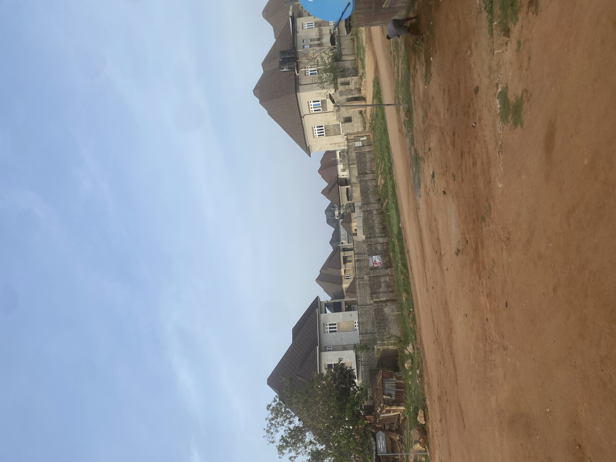 The scene depicts a residential street in the Iddo Tudunwada area of Nigeria. The photo is angled, showing a row of two-story houses along a reddish-brown dirt road. The houses appear to be relatively new, with unfinished concrete walls and dark-colored roofing. Fences line the properties. A few trees are visible, and the sky is pale blue. A person is at the end of the street. Some construction materials are on the ground.