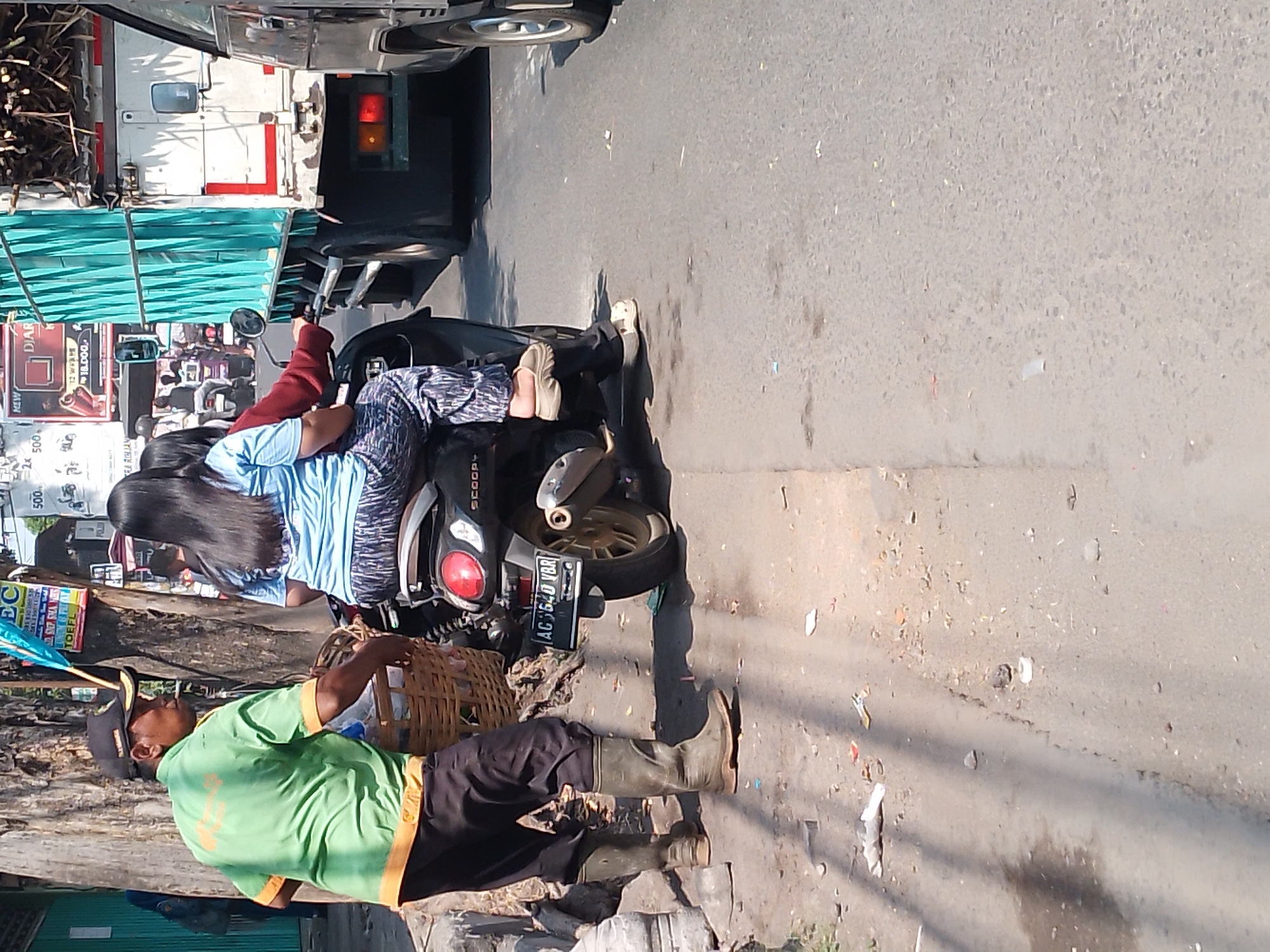 A street scene in Kertosono, Indonesia, captures a man wearing a green shirt and black pants, with tall green rubber boots, holding a woven basket. He stands next to a motorcycle carrying two people. The woman in front, wearing a blue and white striped shirt and patterned pants, is the passenger, while the man behind her wears a red jacket. The motorcycle is parked on the side of a road, with a large white truck with a turquoise tarp cover visible in the background, along with other vehicles. The road is paved and appears to have some debris scattered on it, suggesting it is a public street in a busy area. The lighting indicates it is daytime.