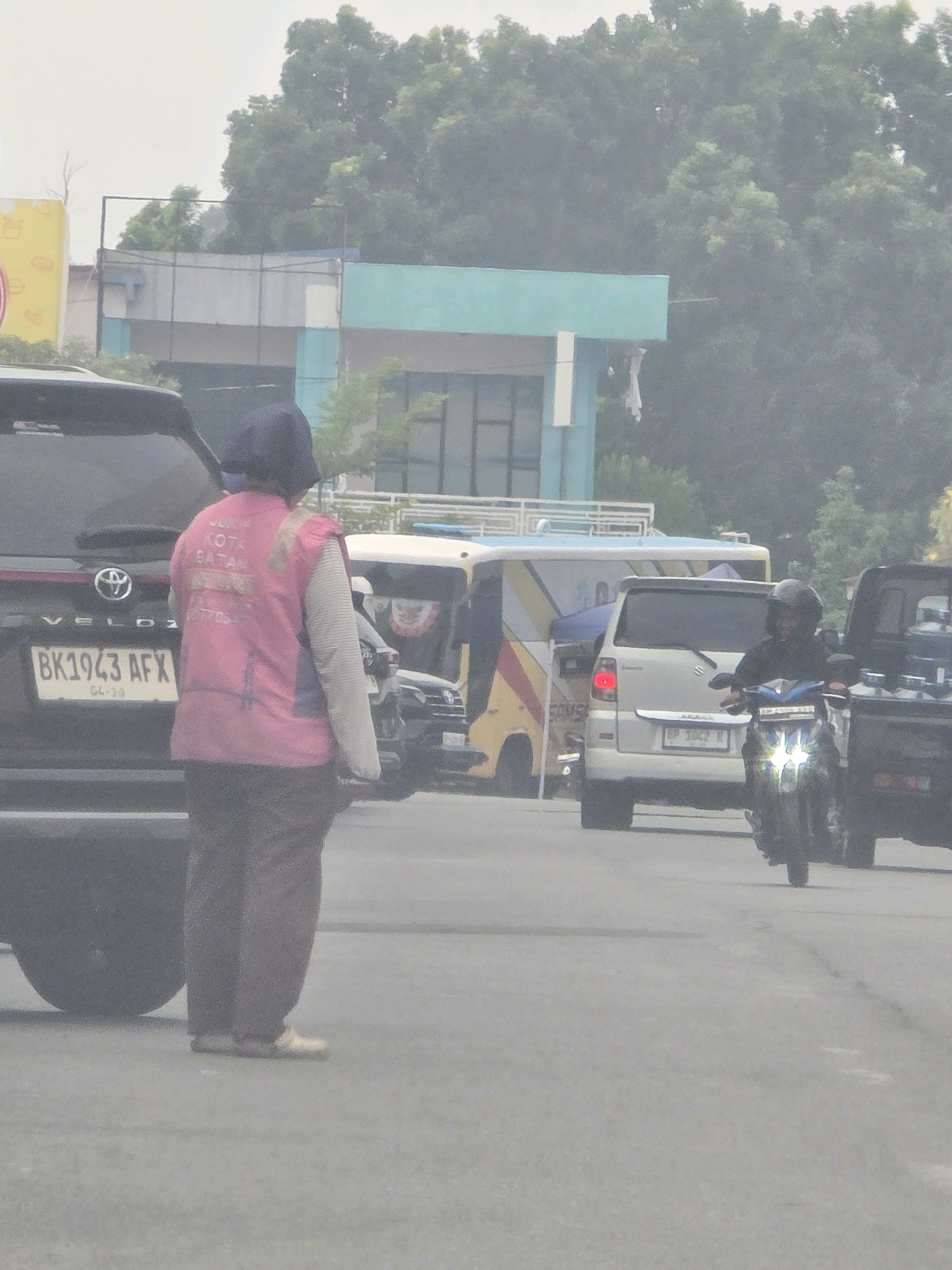 The image shows a street scene in Batam, Indonesia. In the foreground, a person wearing a pink vest over a light-colored shirt and brown pants stands facing away from the camera, looking towards the left. The vest has text on the back, which appears to be in Indonesian. A black SUV is parked to the left of the person, with a license plate that reads "BK1943 AFX". Behind the person and the SUV, several vehicles are visible, including a white van with a yellow and red stripe, a silver car, and a motorcycle. A person on the motorcycle is wearing a helmet. In the background, there are buildings and trees, suggesting an urban environment. The sky is overcast. The overall atmosphere appears to be hazy or smoggy, which is common in industrial areas. The presence of the vehicles and pedestrian indicates a public street, likely in a commercial or residential area of Batam.