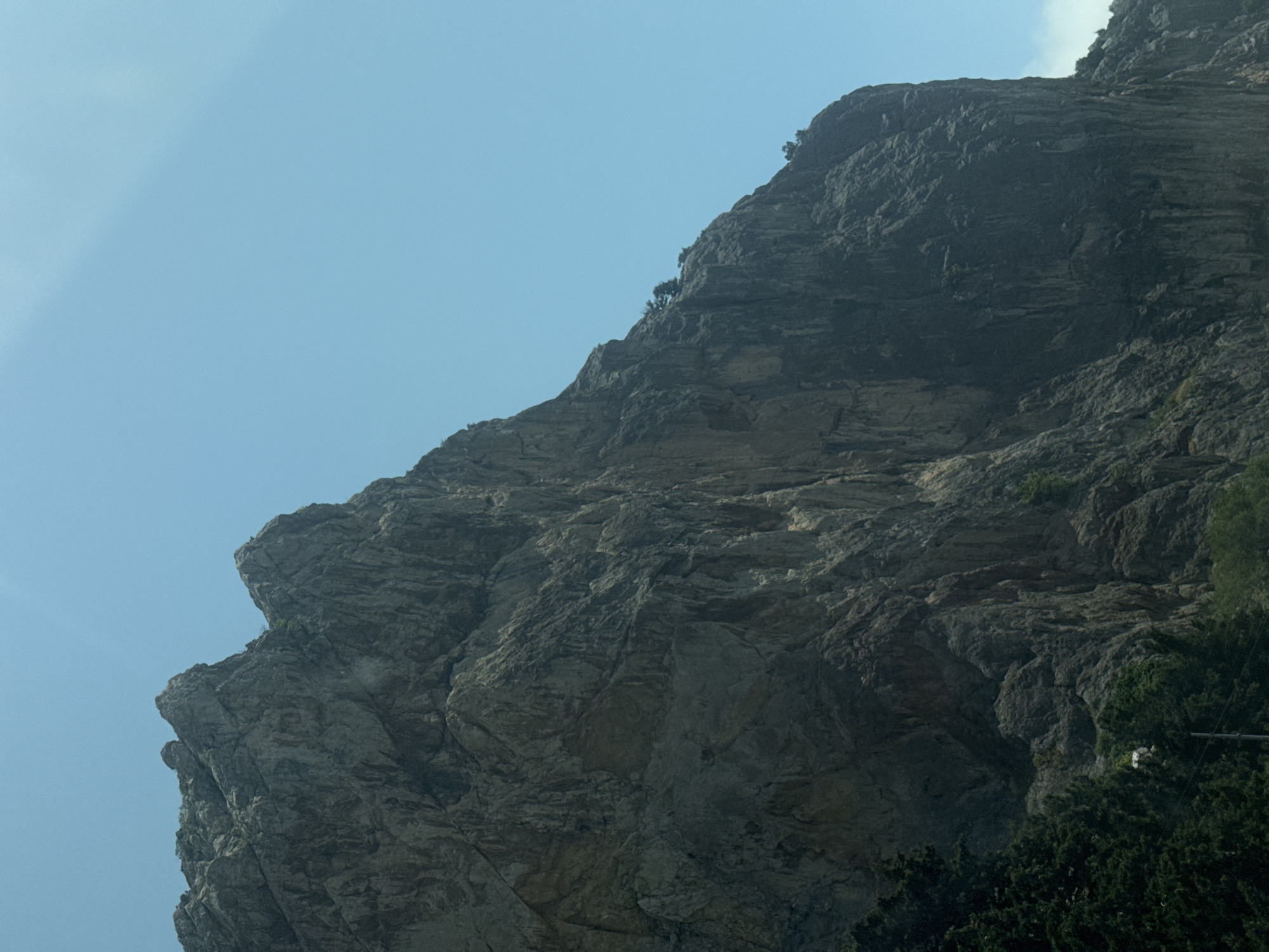 A low-angle shot depicts a mountain against a clear, light blue sky. The mountain occupies most of the frame, exhibiting a rough, rocky surface with various shades of gray. A few small trees cling to the mountainside. Patches of green foliage can be seen at the bottom right of the frame. The location is Орлиновский округ, Ukraine.