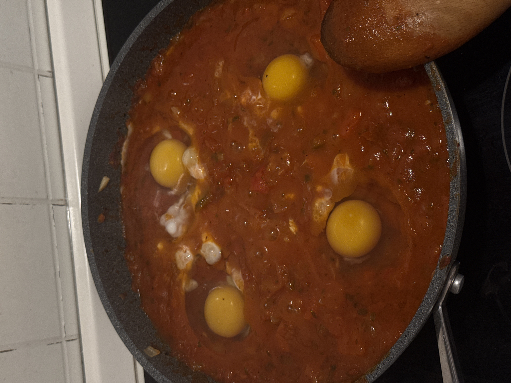 A close-up, high-angle shot captures a dark gray frying pan filled with a vibrant, reddish-orange tomato-based sauce, actively cooking. Scattered within the thick sauce are four distinct, bright yellow egg yolks, with some translucent egg white visible around them, suggesting they have been recently cracked into the pan. The sauce appears to contain small pieces of vegetables or herbs, adding texture and depth to its color.
A wooden spoon is partially visible at the top right of the frame, its handle and part of its bowl resting near or in the sauce, implying it's either just been used for stirring or is awaiting use for serving. On the left side of the image, a section of a white tiled wall with visible grout lines can be seen, providing a domestic kitchen setting. The right side of the pan rests on a dark surface, likely a stovetop.
The activity depicted is the cooking of a dish resembling shakshuka or eggs poached in tomato sauce. The lighting is somewhat dim but focused on the food, suggesting an indoor setting, possibly in the evening. There is no visible text in the image. The location context provided for this scene is Milan, Italy.