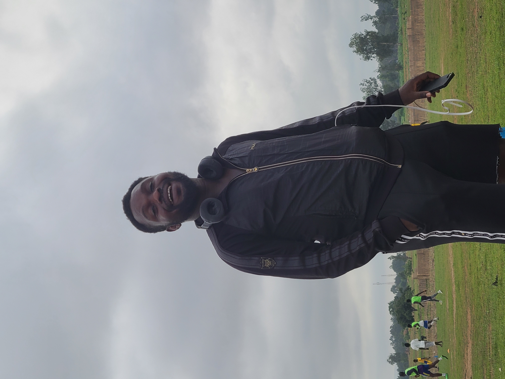 The image features a young man in the foreground, standing on a grassy field under a heavily overcast sky. He is looking directly at the viewer with a broad, genuine smile, revealing his teeth. He has short dark hair and a beard, and is wearing black over-ear headphones around his neck. His attire consists of a dark, possibly black, zip-up athletic jacket with triple stripes on the sleeves and a small emblem on the left chest that appears to read "OSIOS." He also wears dark shorts. His right hand holds a black smartphone, from which white earbud wires hang loosely.
In the background, a wide green field is visible, populated by several other individuals, mostly appearing younger. Some of these individuals are in crouched or starting positions, suggesting they are participating in an athletic event, a training session, or a race. They are dressed in various sportswear, including neon green and white tops. Beyond the field, a line of trees marks the horizon, with some indistinct structures or power lines faintly visible in the far distance under the uniform gray sky. The overall scene depicts an outdoor sports or training gathering on a cloudy day, with the main subject appearing cheerful and relaxed. Visible text includes "OSIOS" on the jacket's emblem and "USA New Balance" faintly on the jacket's interior.