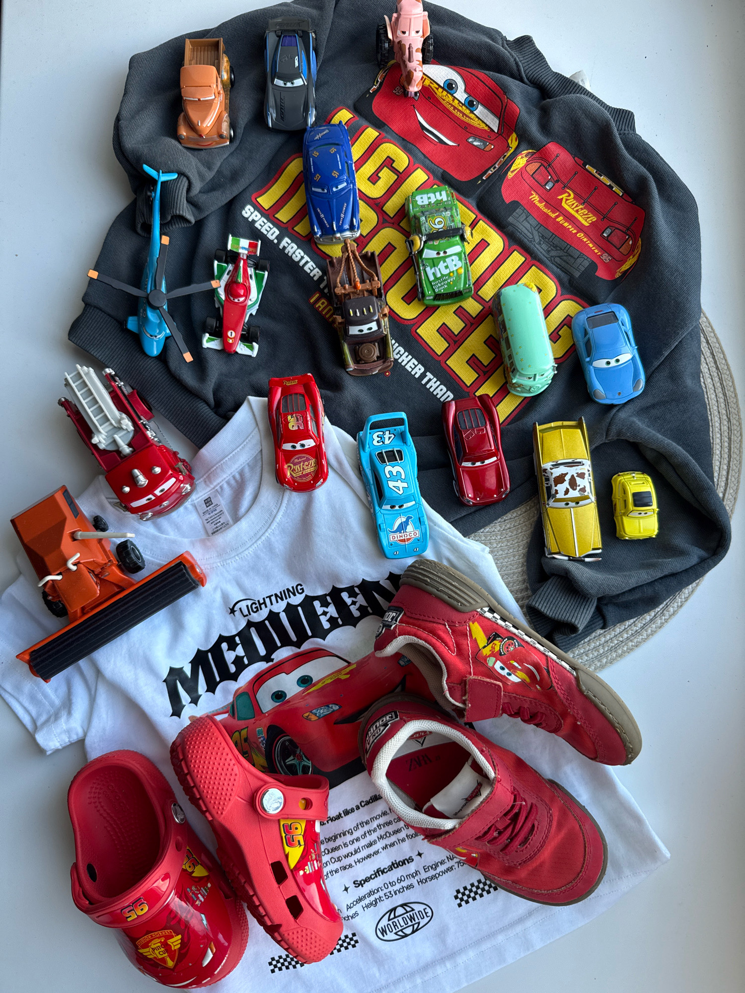 A flat lay of children's clothing and toys, primarily themed around the Disney Pixar Cars franchise, is displayed on a white surface. The collection includes a dark gray sweatshirt with a large graphic of Lightning McQueen, a white t-shirt with a similar graphic and text, a pair of red Crocs with Cars imagery, and red sneakers also decorated with Cars characters. Scattered amongst the clothing are various die-cast toy cars, including a blue helicopter, a red race car, a green race car, a brown tow truck, a blue sedan, a red sedan, a yellow car with a cow print design, a small yellow car, and an orange truck with a ladder. A red toy fire truck is also present. The items are arranged in a somewhat scattered yet organized manner, suggesting they are being presented or displayed. The lighting is even, and there are no visible shadows suggesting an indoor setting. There are no people in the image, and no specific geographical landmarks are identifiable that would place the scene in Rusanovo, Russia.