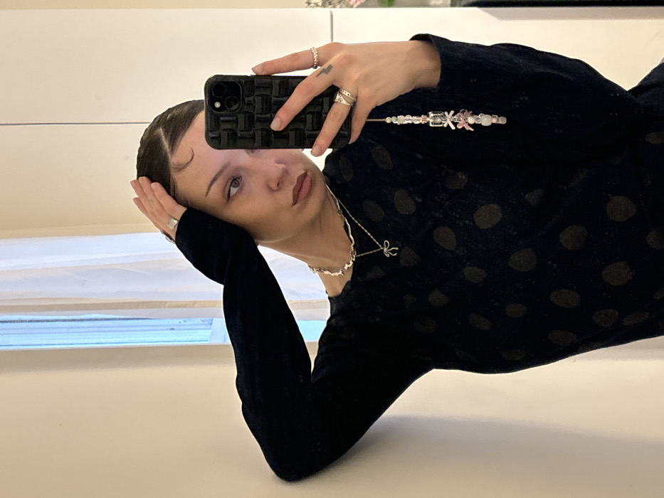 The image displays a woman taking a mirror selfie in an indoor setting. She appears to be in her 20s or 30s, with dark hair styled back, featuring baby hairs on her forehead. She wears a dark, long-sleeved top with a subtle textured or polka-dot pattern. Adorning her neck are two silver necklaces: one a wavy chain and the other a finer chain with a silver bow pendant. Her left hand, holding a black smartphone encased in a textured black cover and accessorized with a multi-colored beaded phone charm, exhibits several rings on her index, middle, and ring fingers, along with a small tattoo on her pinky. Her right hand, featuring a plain silver ring on its index finger, is positioned near her temple, lightly touching her hair. The background consists of a plain, light-colored wall on the left and a window partially covered by sheer white curtains on the right. No specific identifiers linking the scene to a particular location within London, United Kingdom, are discernible.