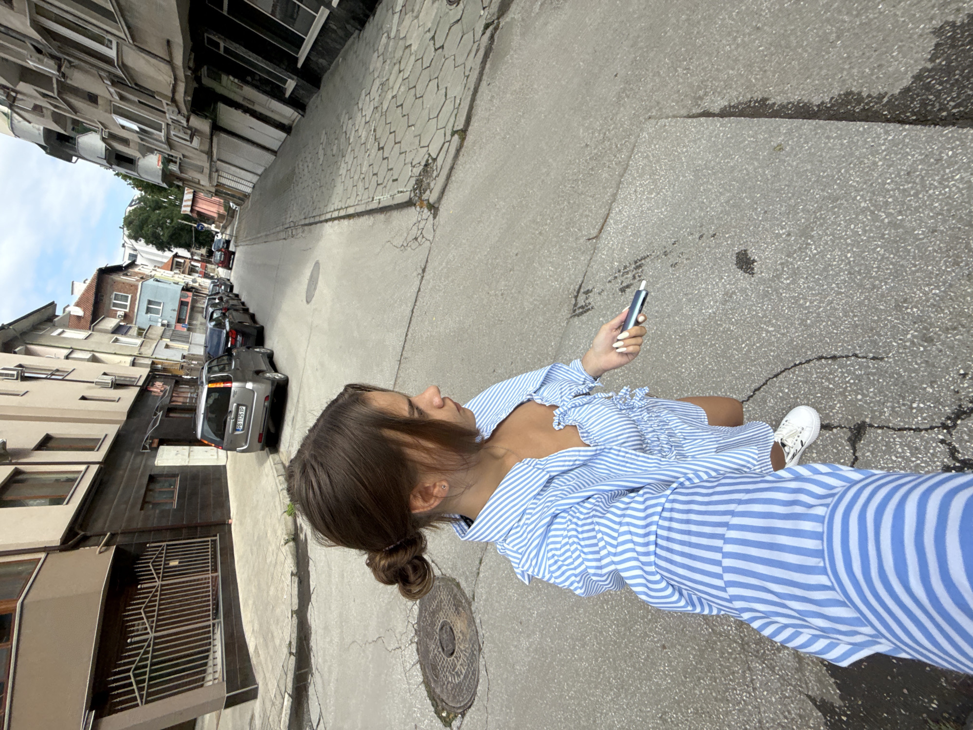 A young woman with brown hair in a bun is captured from a low-angle perspective, holding a silver device in her right hand. She is wearing a blue and white striped shirt and white sneakers. The woman is standing on a cracked asphalt street in what appears to be an urban setting. In the background, buildings with various architectural styles line the street, and several cars are parked along the side. The street has a cobblestone sidewalk on the right. The image quality is decent, with good lighting and clarity, suggesting it was taken during the daytime. The background elements suggest a European city, possibly Plovdiv, Bulgaria, given the architectural style of the buildings and the presence of parked cars.