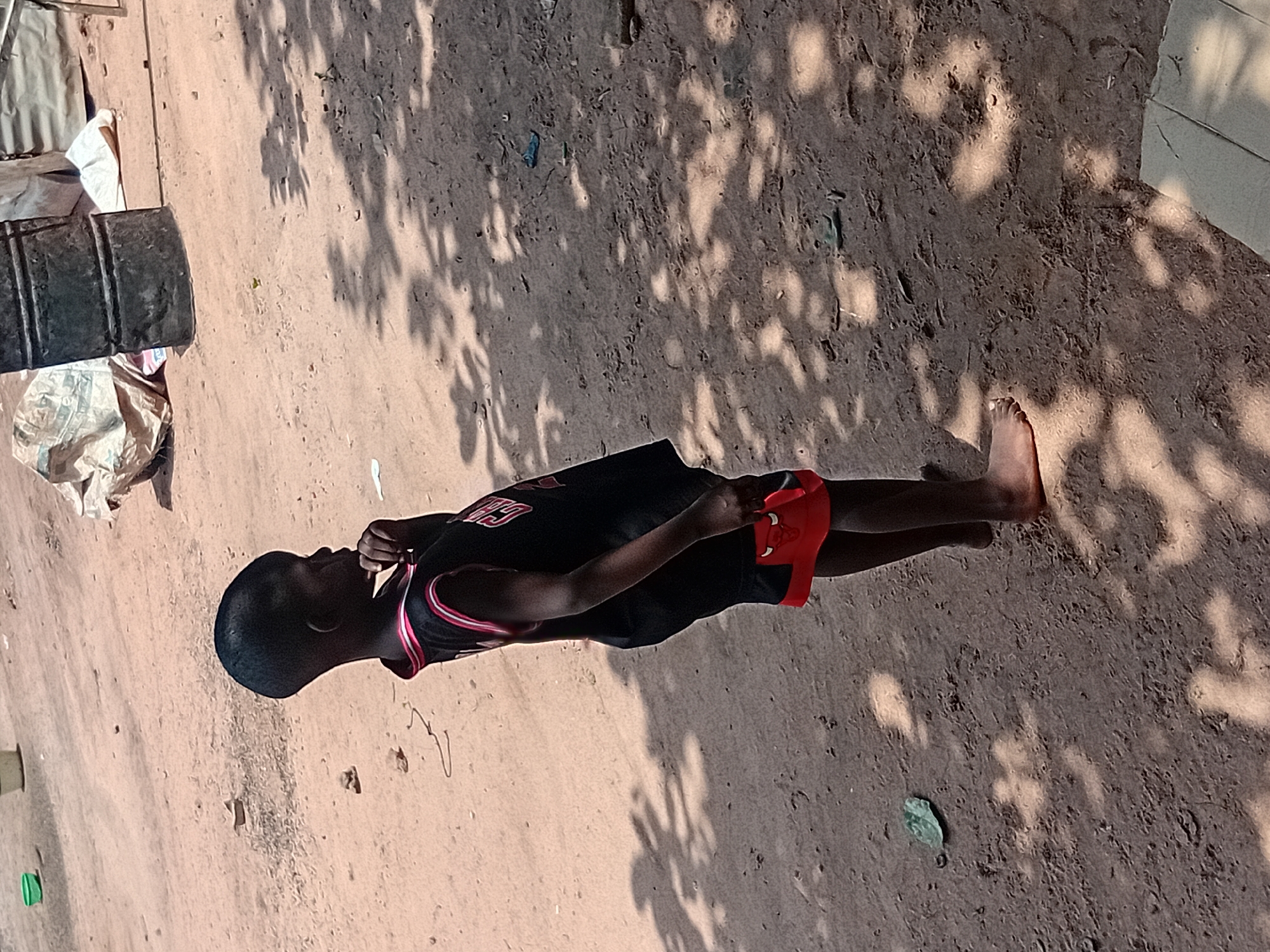 A young boy stands on a dirt ground in the city of Ejule, Nigeria. He is wearing a black tank top with red and white trim and red shorts with a red bull logo. The boy is facing to the right and has his right hand to his chin, as if in thought. The ground is covered in loose dirt and small stones, with scattered leaves and shadows from trees overhead. In the background, there are several objects including a large black barrel, bags of materials, and some corrugated metal structures. The overall scene appears to be an outdoor area, possibly a yard or open space in a residential or commercial area.