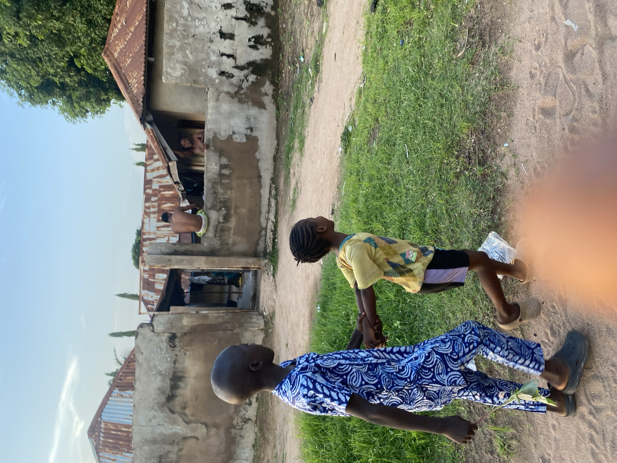 In Jauro Yinu, Nigeria, two young children walk hand-in-hand on a dirt path lined with green grass. The child on the left wears a blue and white patterned tunic and pants, with dark clogs on their feet. They are holding a leafy green plant in their left hand. The child on the right wears a yellow t-shirt with a geometric pattern and dark shorts with a purple stripe, and light-colored clogs. In the background, there are several structures with corrugated metal roofs and rough, light-colored walls. Two men are visible on top of a wall in the mid-ground; one is shirtless, sitting with his legs dangling, and the other is leaning over the wall, looking towards the viewer. The sky is clear blue with some wispy white clouds.