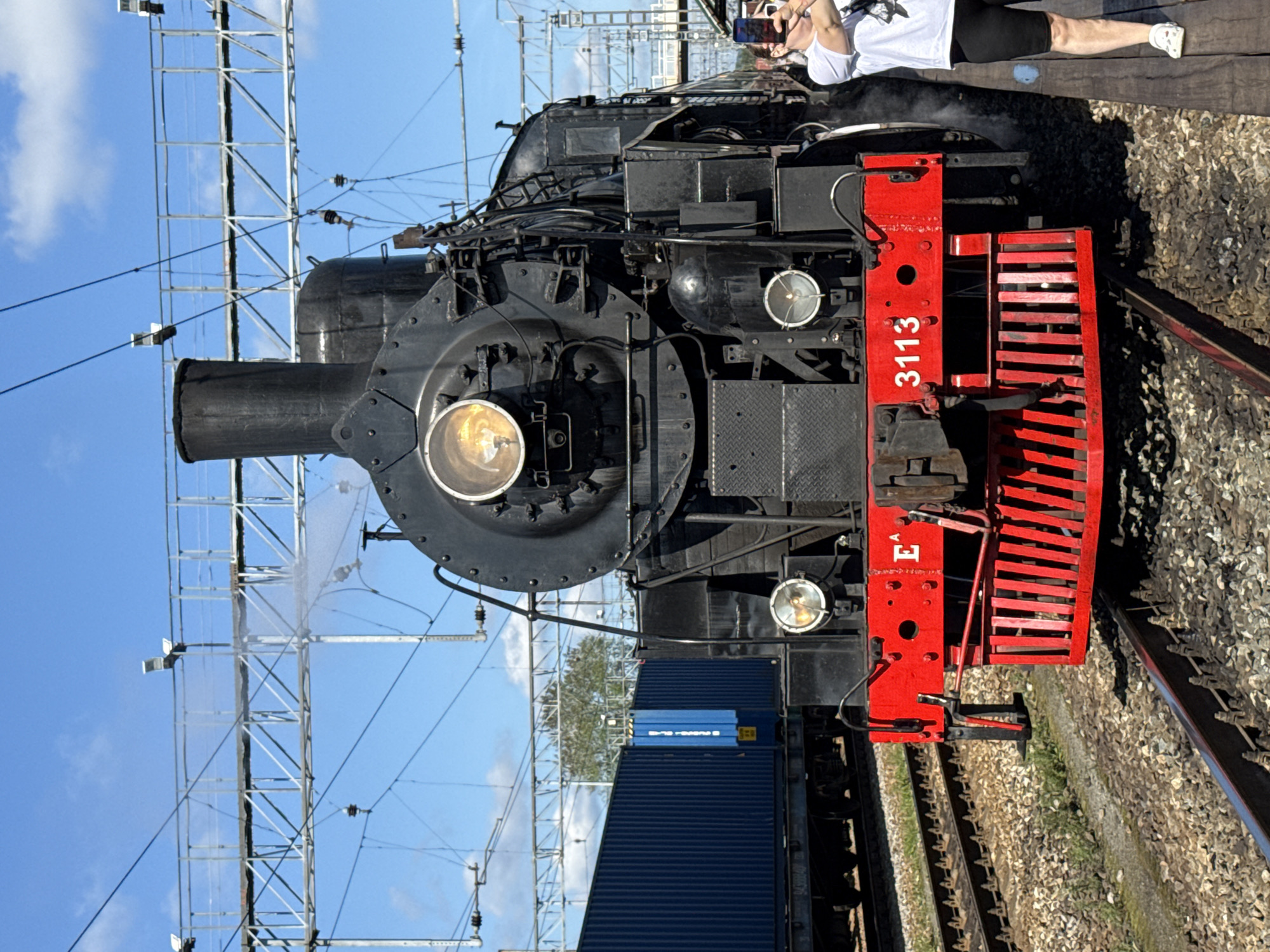 This is a medium shot, captured in Yekaterinburg, Russia, featuring a vintage black steam locomotive positioned on a railway track. The locomotive is the primary subject, with its prominent headlight illuminated and a plume of steam rising from its chimney. The front of the locomotive is adorned with a bright red buffer beam and a decorative grill. On the left side of the buffer beam, the letter "E" is visible, and on the right, the number "3113" is displayed.
To the left of the steam locomotive, a train car laden with tall, dark blue shipping containers is visible. The background consists of a bright blue sky with scattered clouds and a complex network of overhead electrical wires and support structures.
On the right side of the frame, a woman wearing a white t-shirt and black shorts is standing on a wooden platform. She is holding a smartphone in her hands and appears to be taking a photograph or video of the steam locomotive. The woman is positioned on the edge of the platform, close to the train tracks. The ground beneath the tracks is covered in ballast, with some patches of green grass growing through.