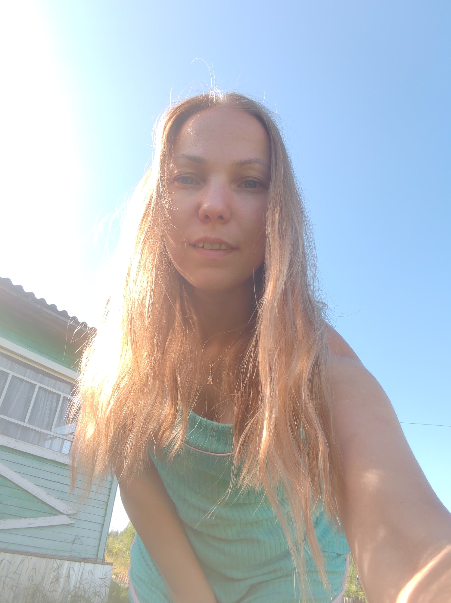This is a low-angle, close-up selfie of a young adult female with long, blonde hair, taken outdoors under a clear blue sky. She is wearing a light blue, ribbed tank top with thin pink trim. A delicate gold necklace with a small pendant is visible around her neck. Her face is partially illuminated by the sun, which is positioned to her left, creating a bright glare. She is looking towards the camera with a slight smile. To the left of the frame, a light blue wooden house with white trim and a corrugated metal roof is partially visible. The house has a window with white patterned curtains. The background suggests a rural or suburban setting, with some greenery and vegetation visible in the lower right corner. The image appears to have been taken in Чухлэм, Russia, likely during daylight hours given the bright sunshine.